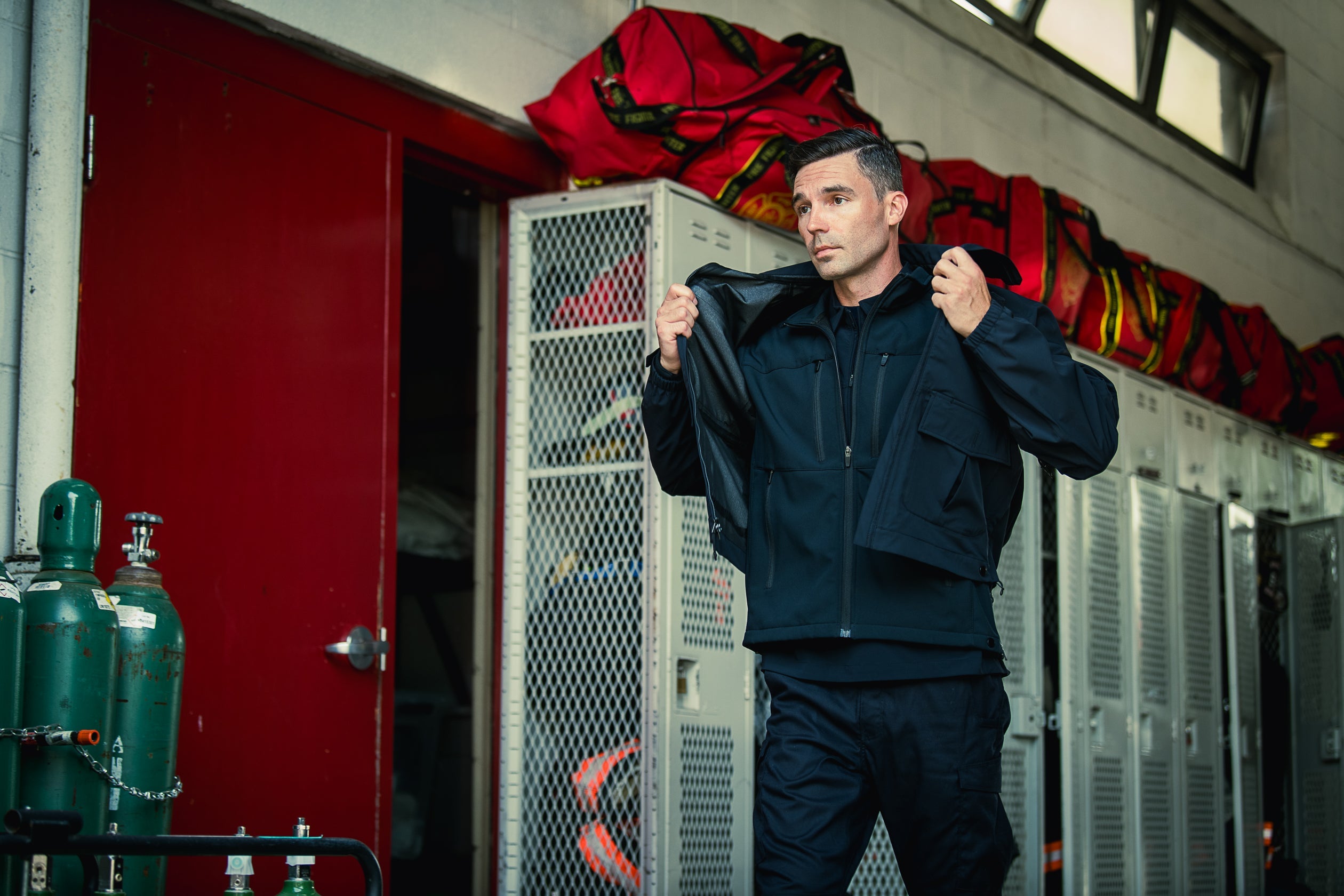 Photograph a man in a dark jacket in a fire station locker room with red doors, metal lockers, and bright red fire equipment bags