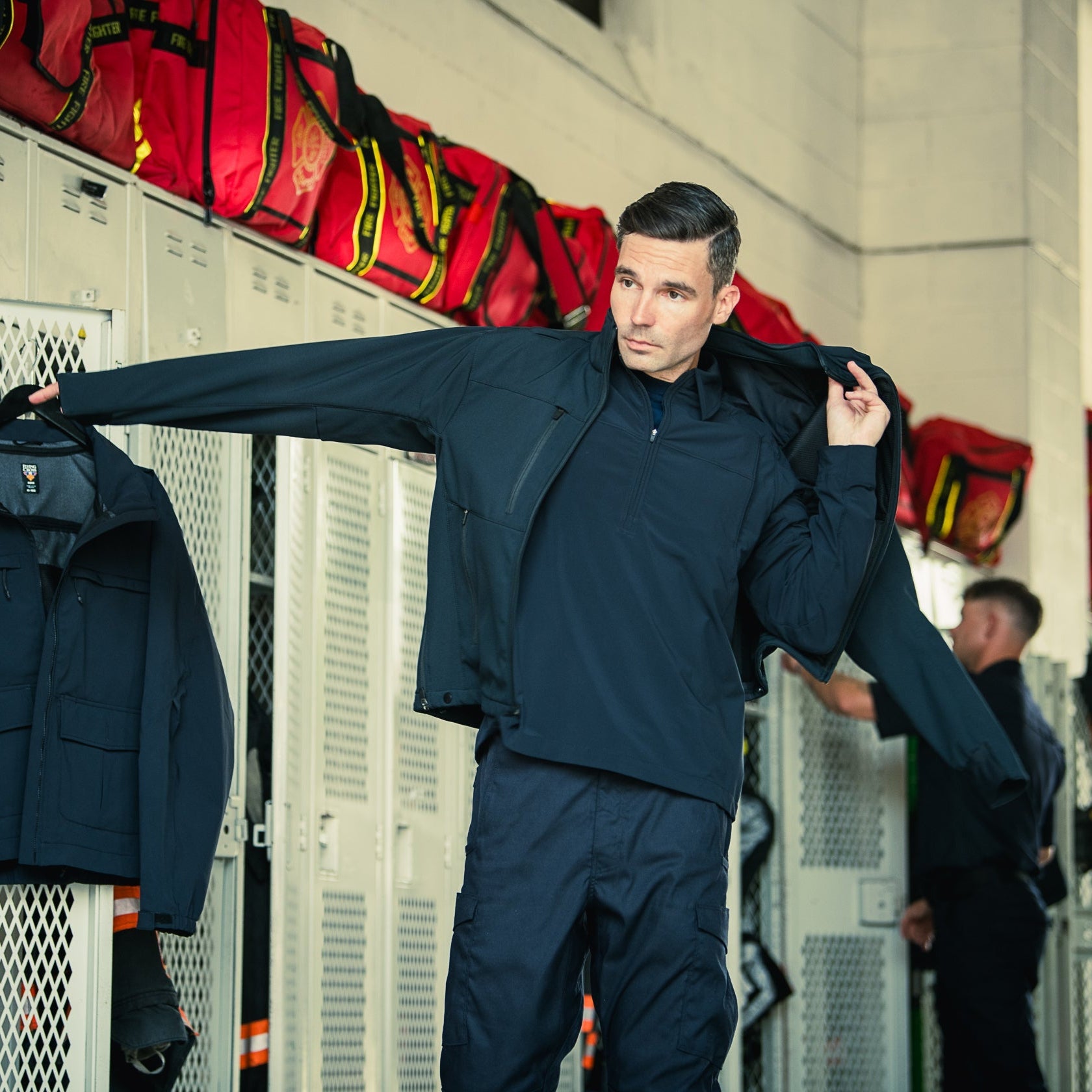 Photography showing a man in a dark navy jacket and pants in a fire station locker room with red fire bags and metal lockers visible along with a partially visible second man and hints of orange safety gear