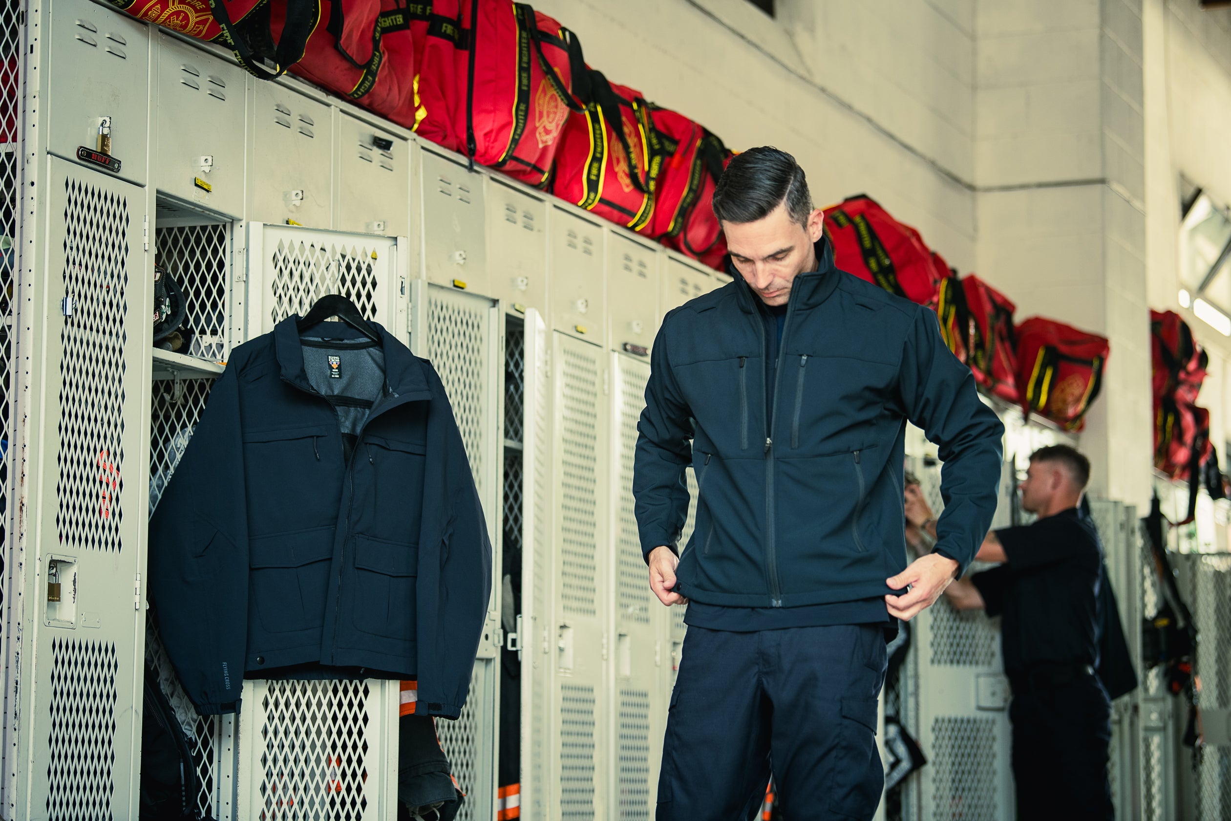 Photograph showing a man in a navy blue jacket in a fire station locker room with red firefighter bags above metal lockers and a dark jacket hanging on a mesh door showcasing its details