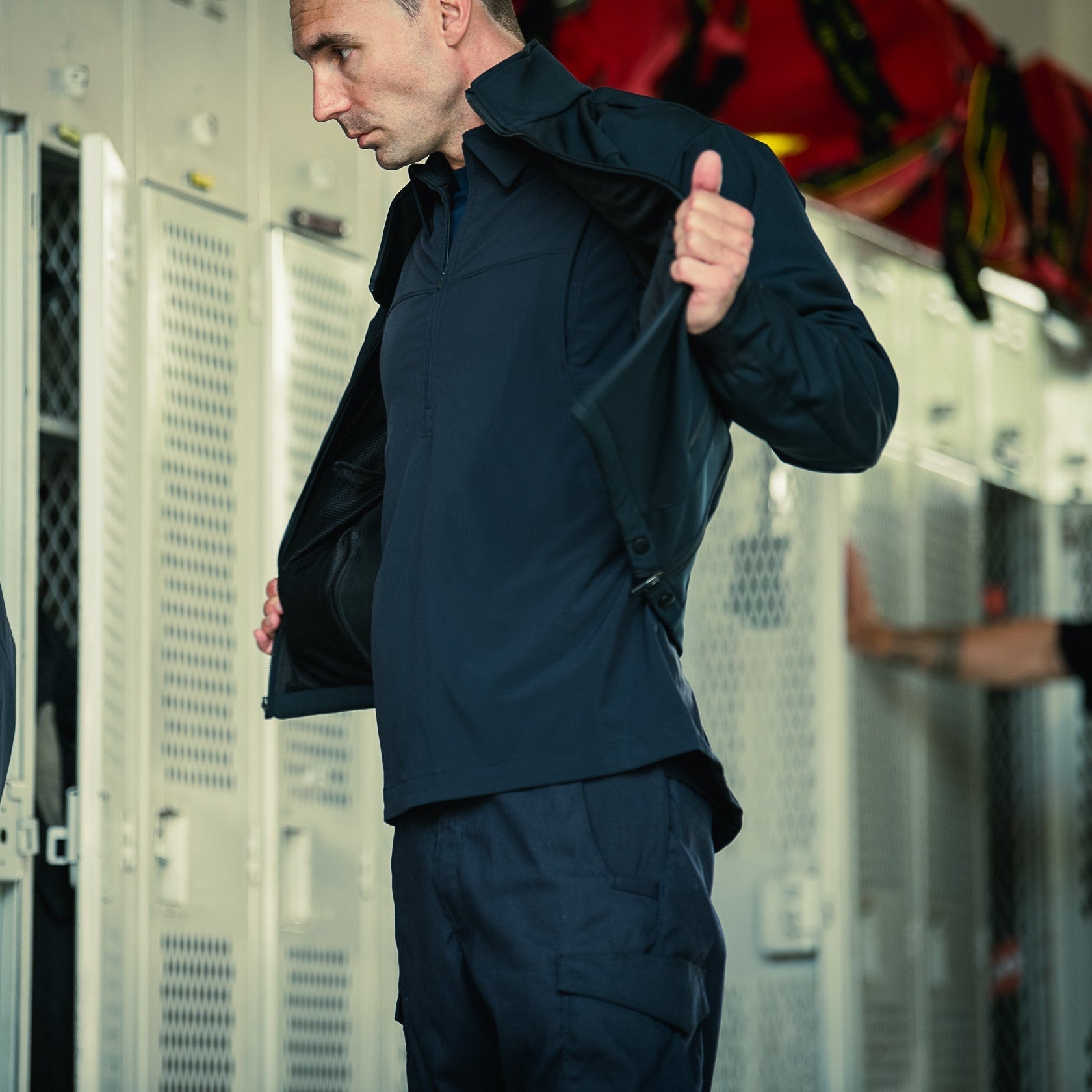 A photograph showing a man in a dark blue tactical jacket and pants in a locker room with metal mesh doors and a red bag in the background, the jacket's inner mesh lining and snap closures are visible
