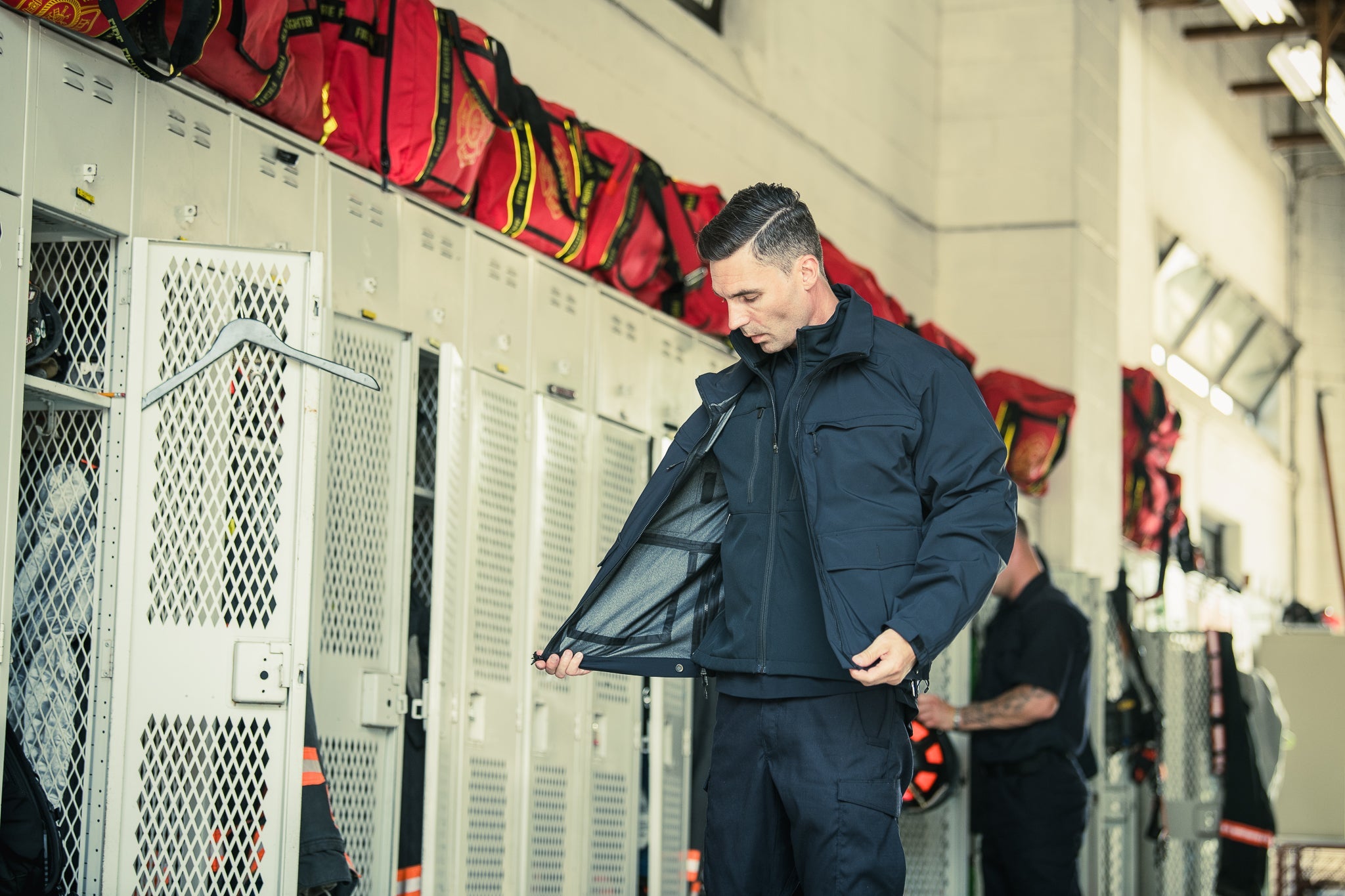 Photograph showing a firefighter in a dark navy jacket with a grey inner lining in a fire station locker room with bright red fire equipment bags and metal lockers