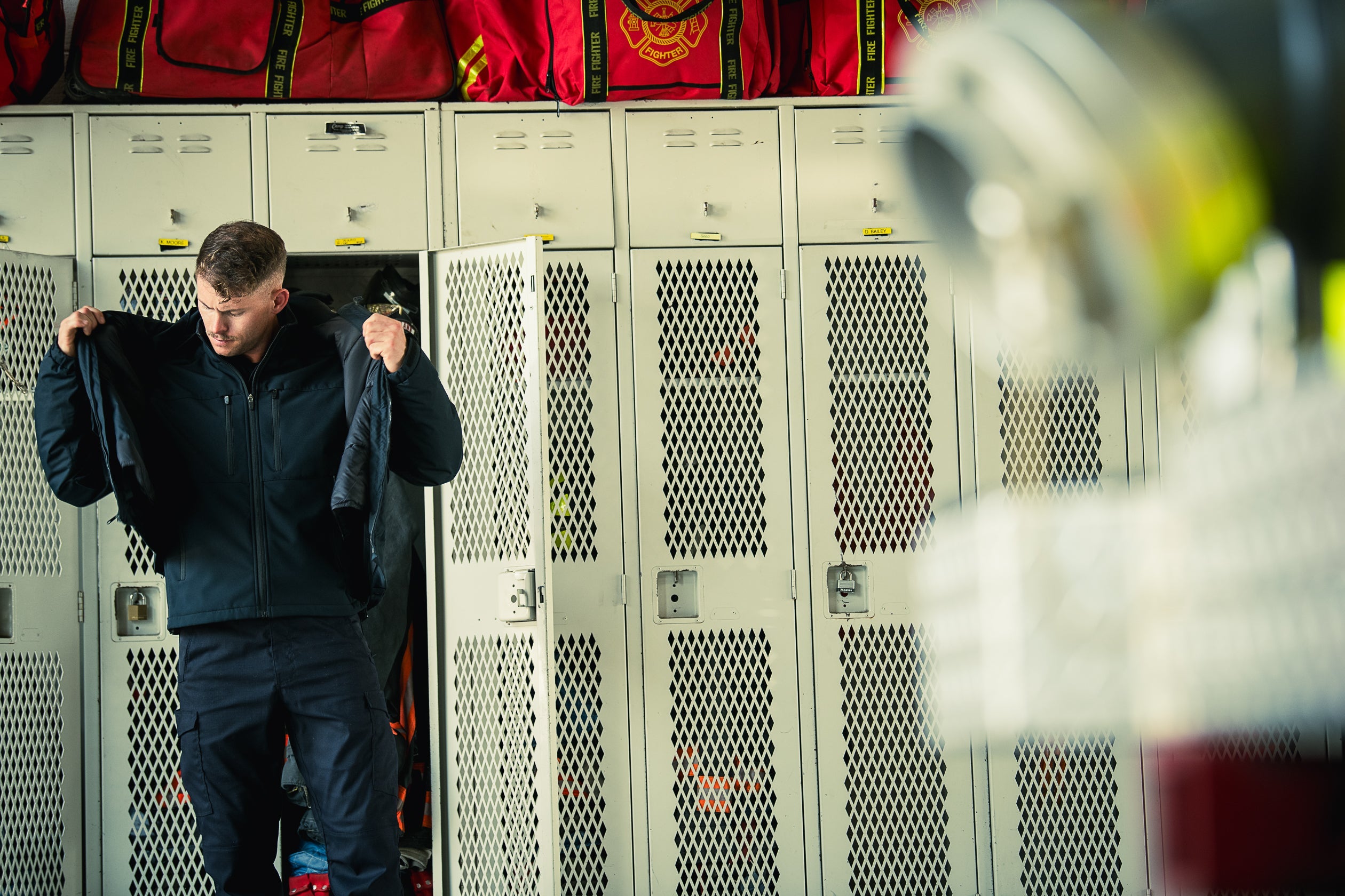 Photograph of a firefighter in a fire station locker room putting on his jacket, near red fire bags and off-white metal lockers with diamond-patterned ventilation, showcasing a brass padlock and name tags