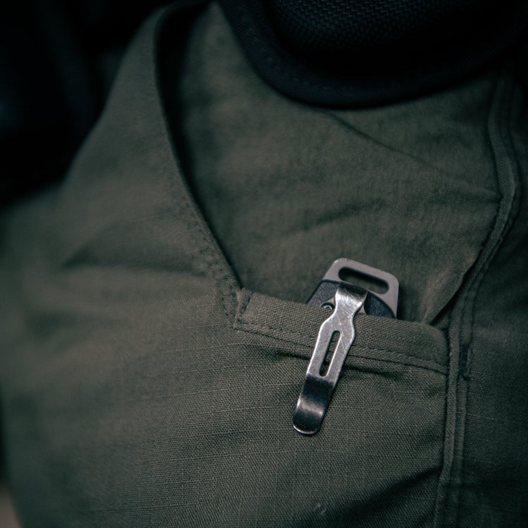Close-up photograph showing a dark gray metallic pocket knife with a unique clip nestled in an olive-green shirt pocket the fabric shows subtle textural details and the knife's metallic finish contrasts beautifully