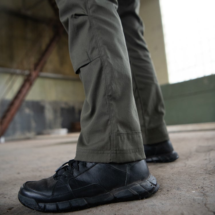 Photograph showing olive green tactical pants and black tactical boots in a dilapidated industrial building with exposed rusted metal beams and a textured concrete floor, showcasing durable fabric, heavy-duty stitching, and a chunky sole