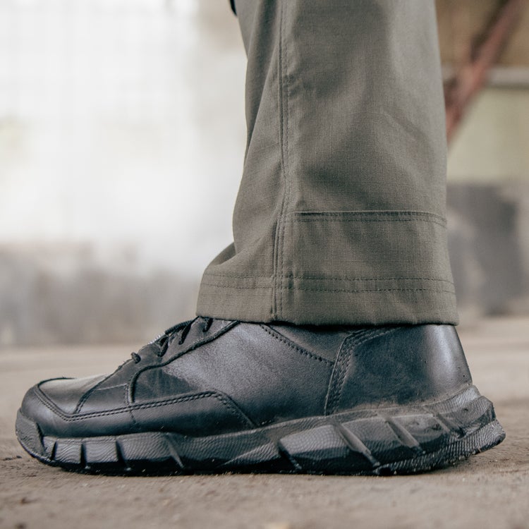 Close-up product shot showing a black leather tactical boot with aggressive tread and olive green pants featuring reinforced stitching detail on the hem
