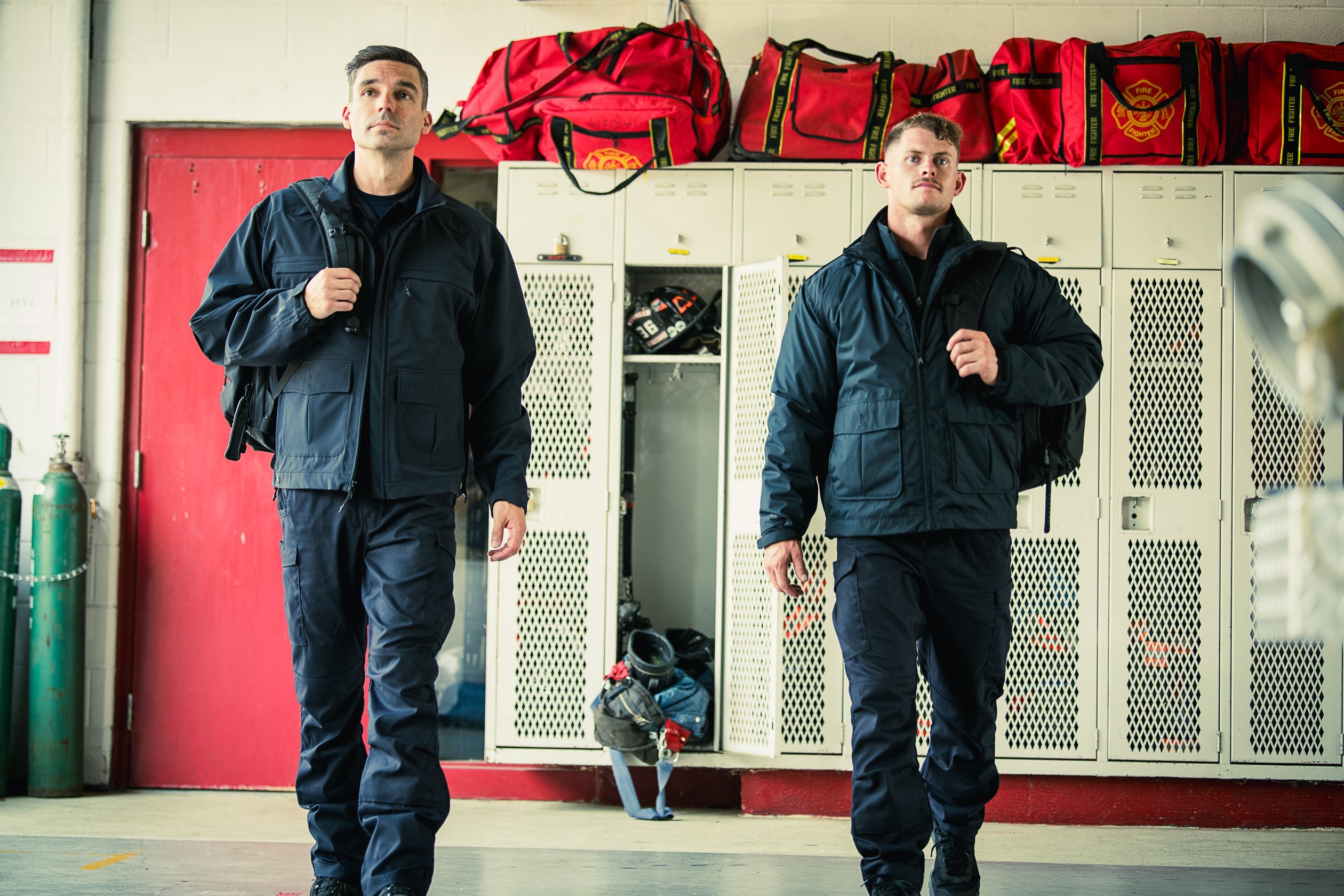 Photograph showing two firefighters in dark uniforms and backpacks walking past lockers and red fire equipment bags in a fire station with a red door and green gas cylinders visible in the background