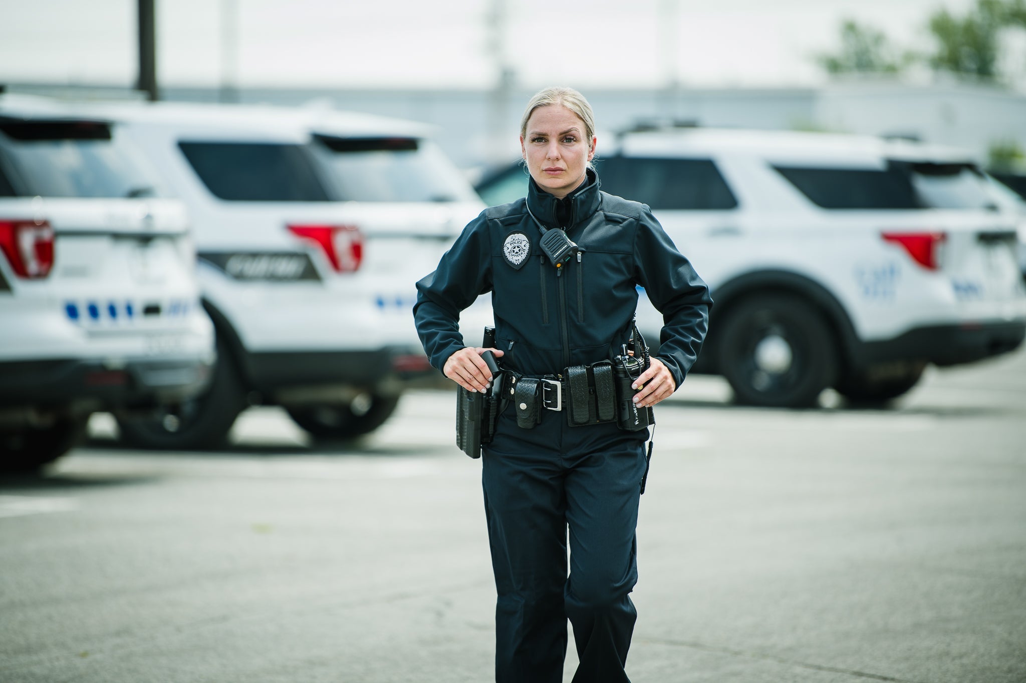Photograph of a female police officer in a dark uniform and duty belt walking in a parking lot with several police SUVs in the background showcasing her badge a radio and a firearm in their holsters