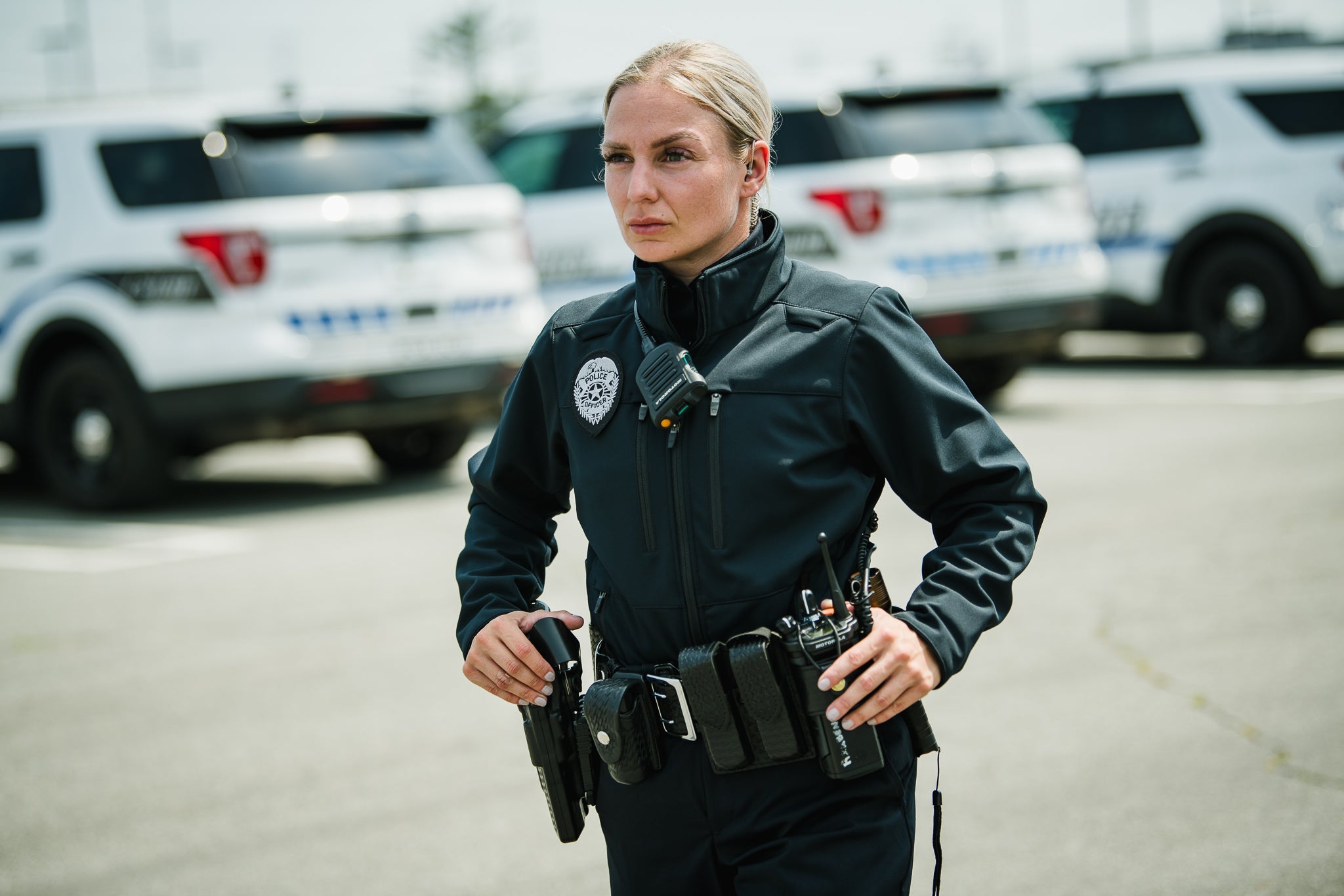 Photograph A female police officer stands in a parking lot with several police vehicles in the background, her dark uniform accented by a silver badge, a black Motorola radio, and a tactical belt