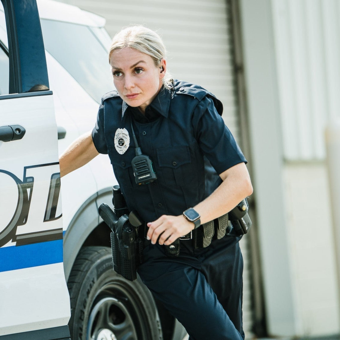 Photograph of a female police officer in a dark-blue uniform exiting a police vehicle near a light-grey building with a Motorola radio a black handgun and a smartwatch visible on her person
