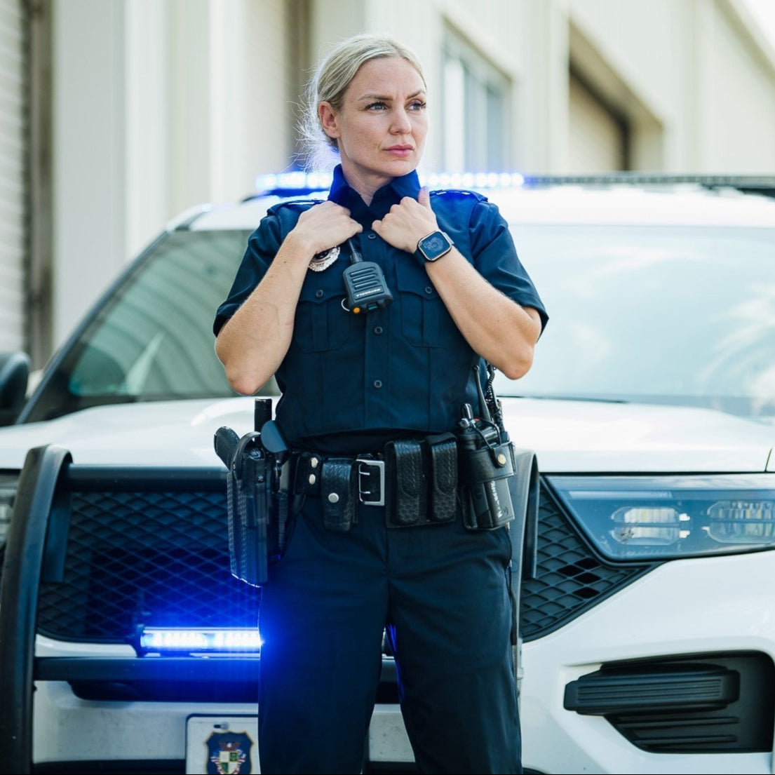 Photograph female police officer in uniform standing before a police car with flashing blue lights, city street backdrop, black utility belt, smartwatch, and radio