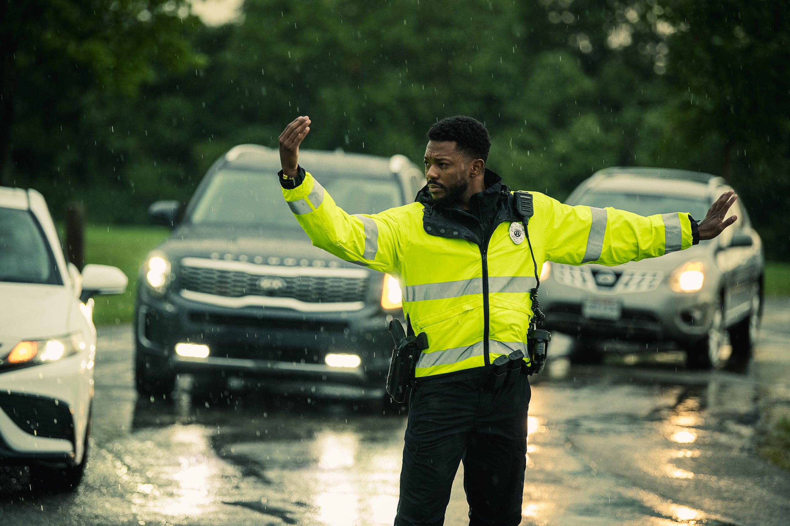 Photograph a police officer directing traffic in the rain near a Kia and Nissan, wearing a bright yellow jacket and amidst glistening puddles and blurry green trees