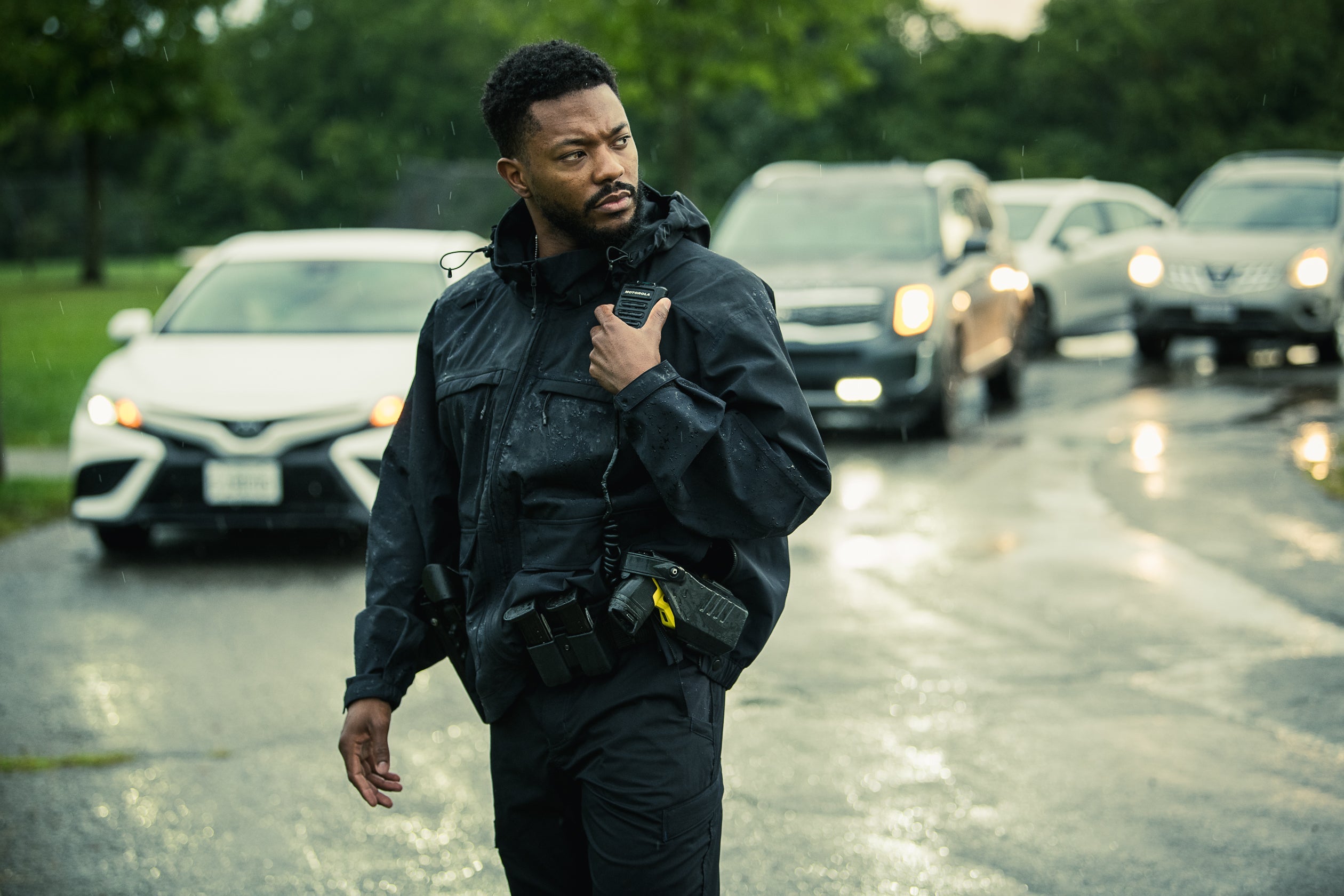 Photograph a uniformed man holding a Motorola radio walks near a rain-slicked road with parked cars and lush green trees in the background, showcasing his dark tactical gear and a yellow detail on his utility belt