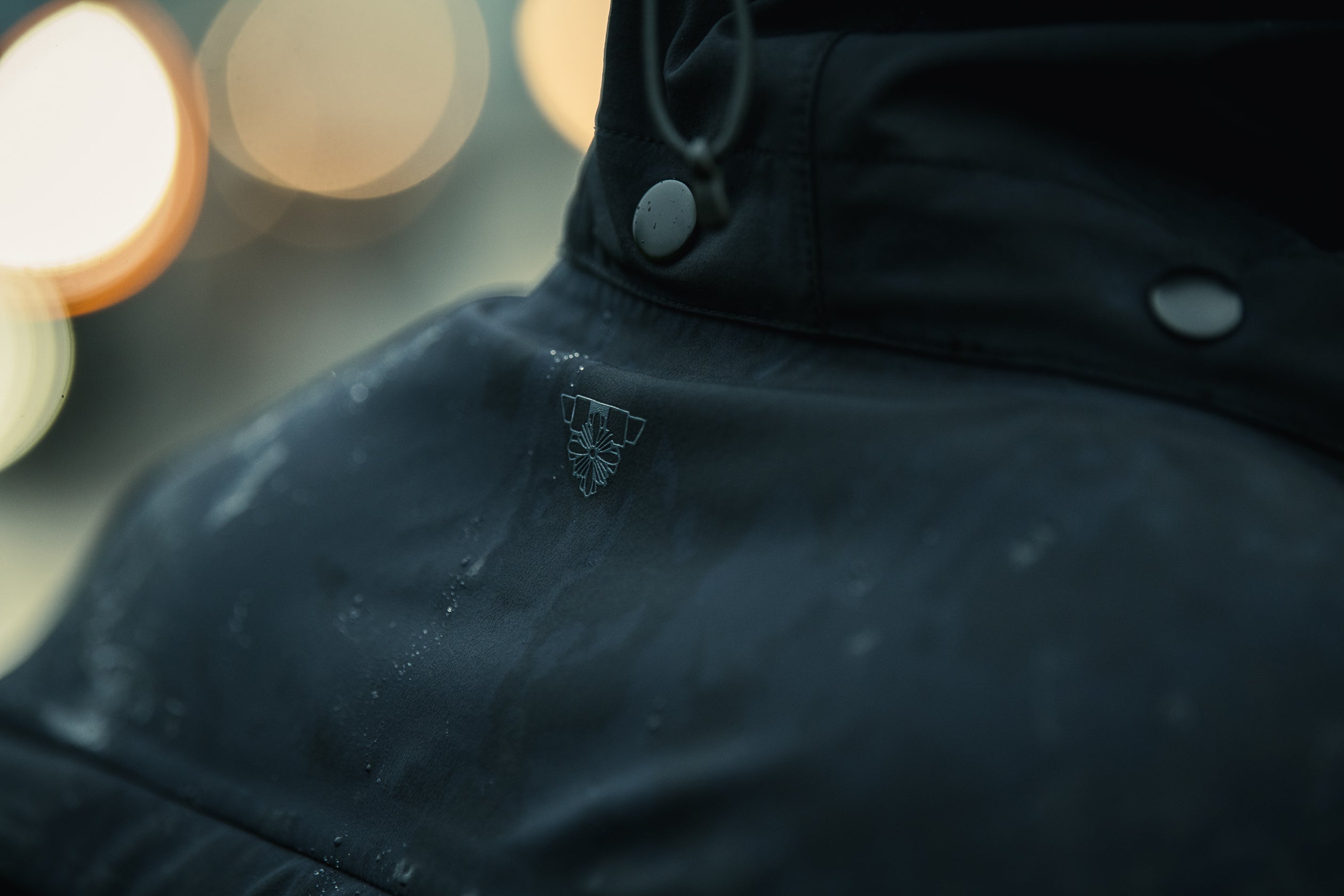 Close-up photograph of a dark teal jacket with a unique embroidered logo featuring a stylized flower, showcasing water droplets and gray snap buttons against a blurred city lights background
