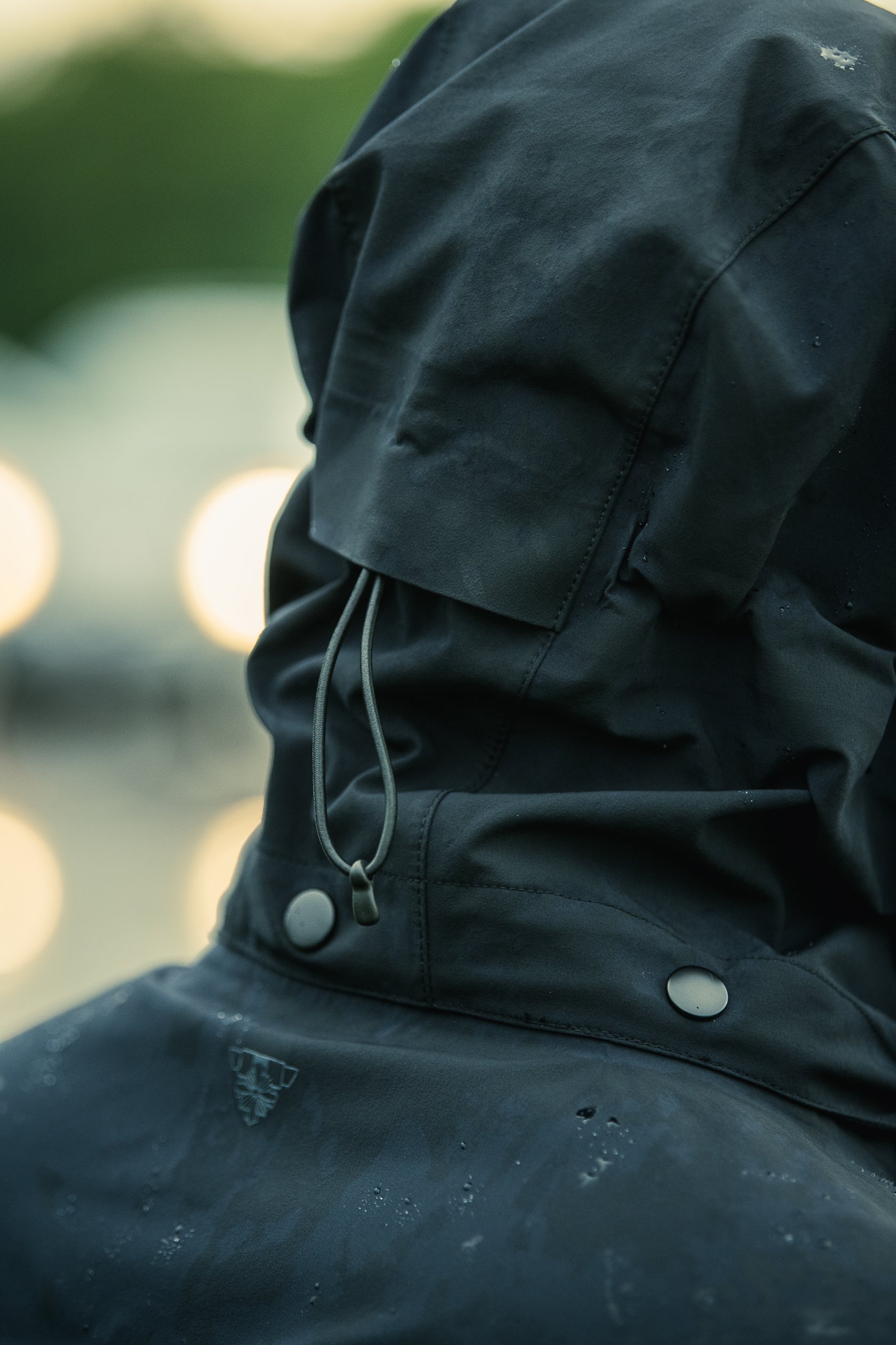 Photograph detail of a dark teal hooded jacket with a drawstring and snap closures showing water droplets and a logo against a blurred background of lights