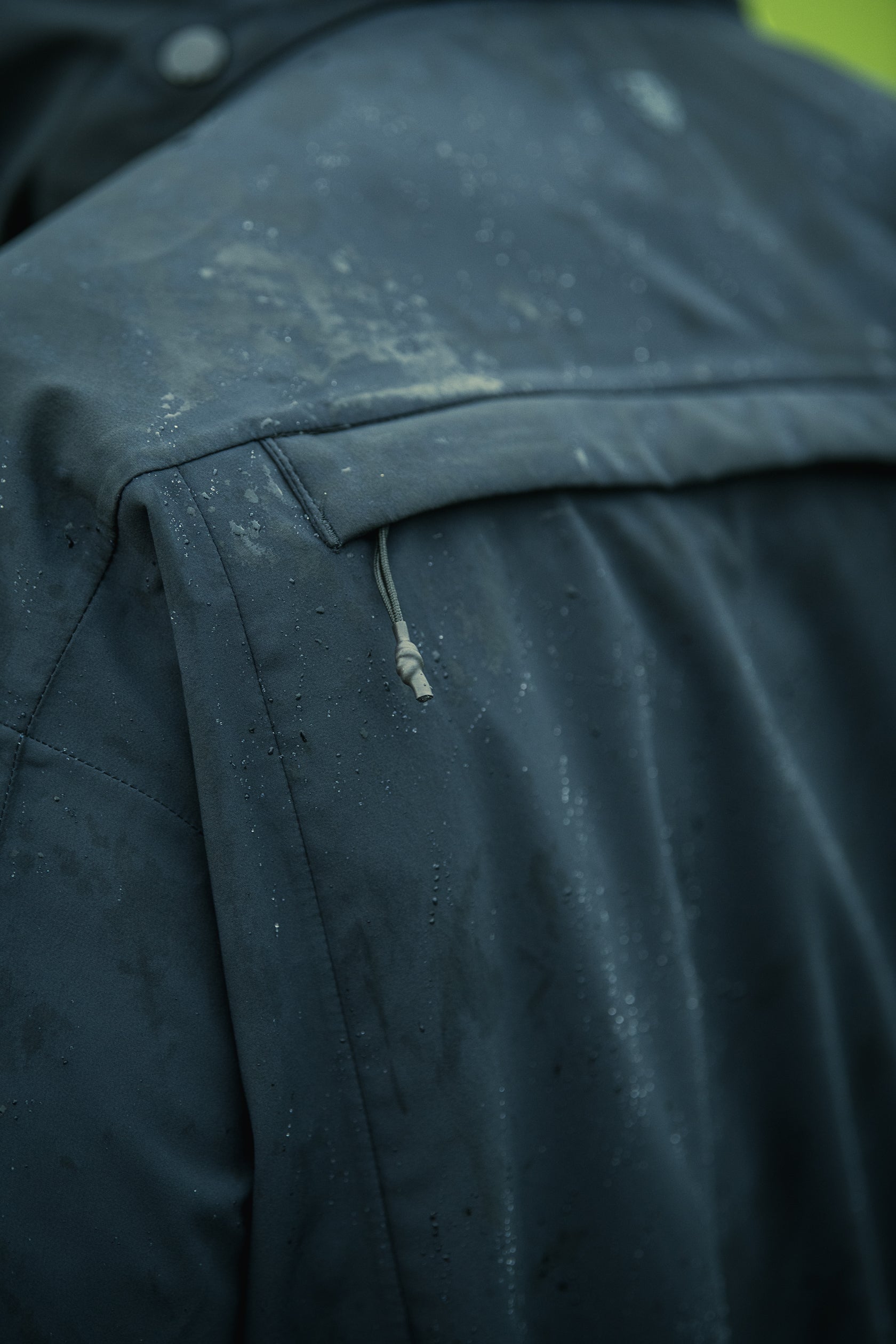 Close-up photograph showing a dark teal waterproof jacket with water droplets, light dirt smudges, and a visible drawstring detail