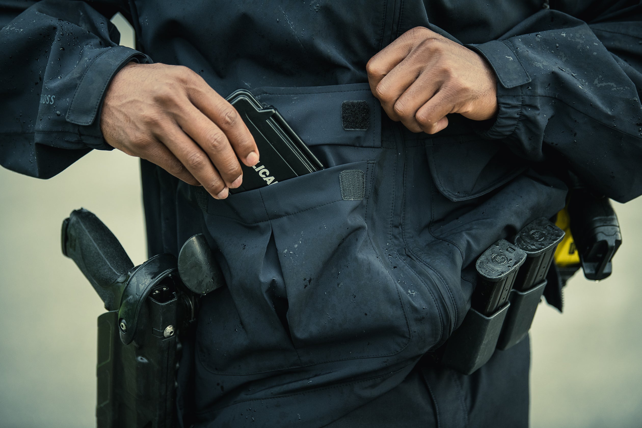 A photograph showing a person in a dark, water-resistant jacket placing a black tactical case into a pocket, with a visible handgun and magazine holders on their hip, and water droplets on the fabric