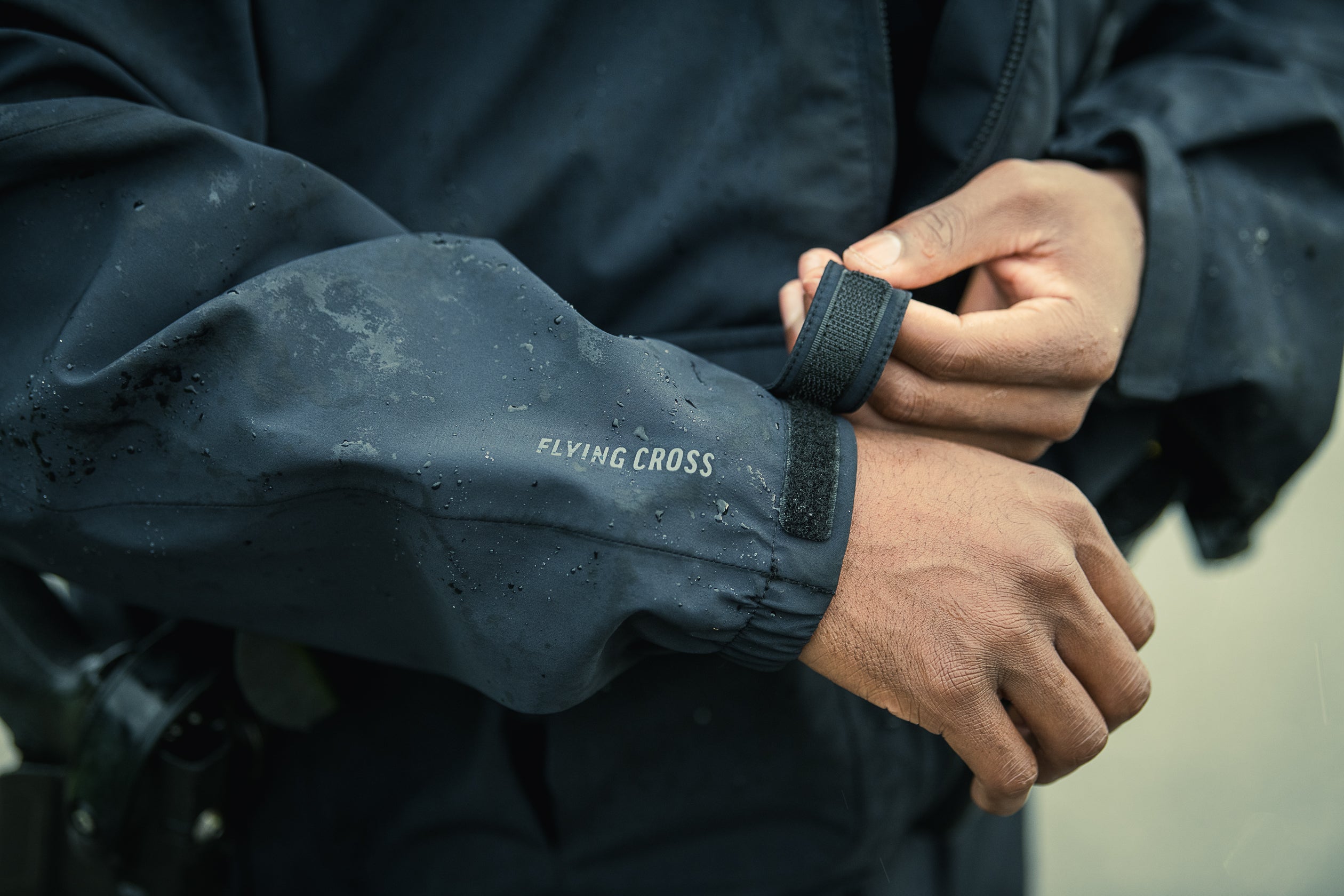 Close-up photo shows a person adjusting the Velcro cuff of a dark, water-resistant Flying Cross jacket showing water droplets and subtle dirt smudges