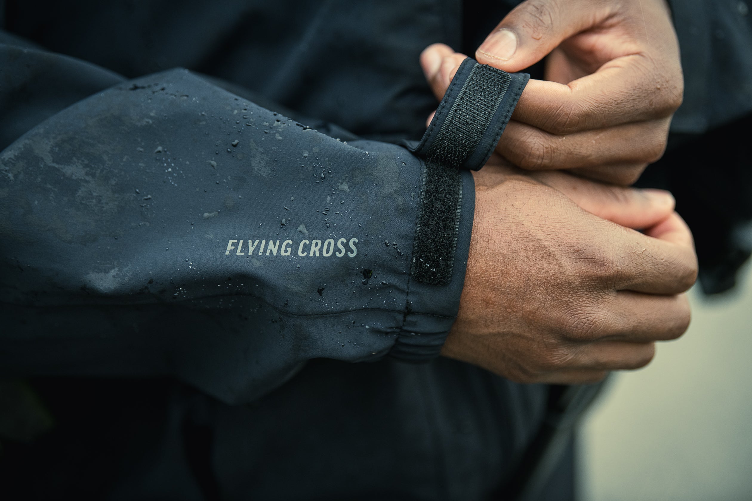 Close-up photograph showing a person adjusting the Velcro cuff of a dark gray Flying Cross rain jacket with water droplets and subtle dirt stains visible on the fabric