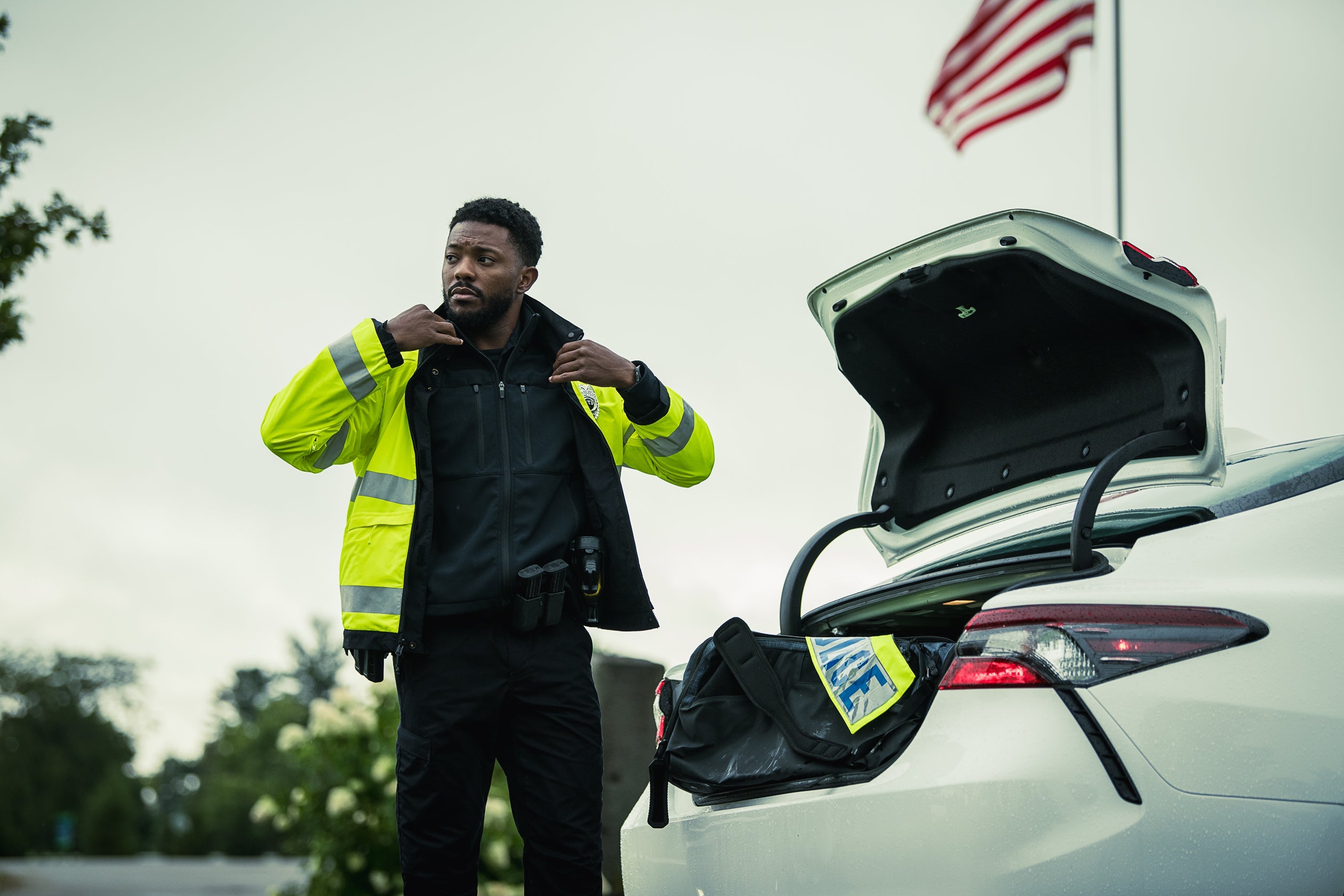 Photograph of a police officer in a bright yellow jacket, near a white car with its trunk open, against a backdrop of trees and a waving American flag, showcasing the officer's serious expression, the reflective strips on his jacket, and the police-marked bag in the vehicle's trunk