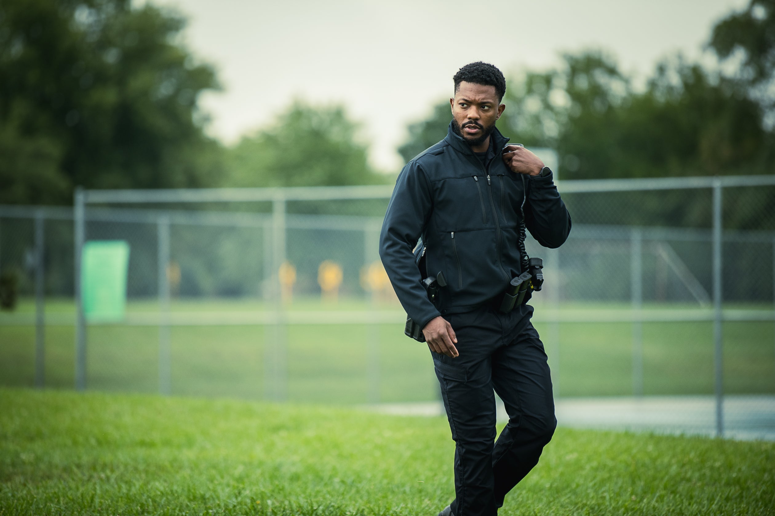 Photograph of a security officer walking on a grassy field near a chain link fence with blurred trees and a partially visible green structure in the background, his dark jacket and tactical gear stand out against the vibrant green and overcast sky