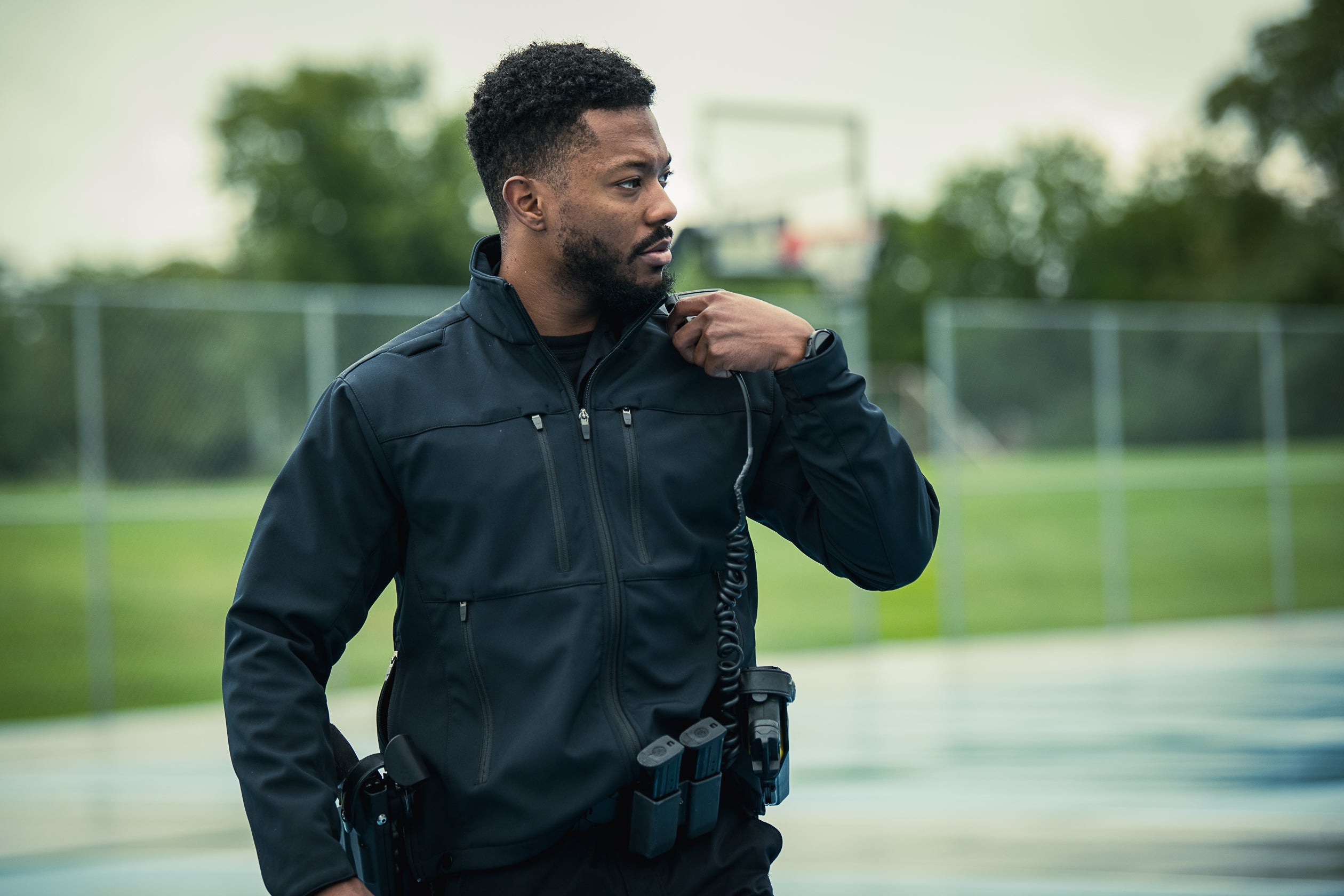 Photograph of a police officer in a dark jacket at an outdoor sports complex with chain link fence and blurred basketball court in the background, featuring a coiled radio cord, a dark watch, and his short curly hair