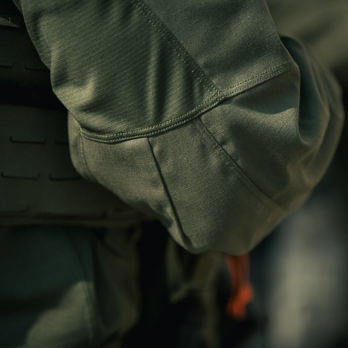 Close-up photograph showing the textured olive-green fabric of a tactical shirt with detailed stitching and subtle shadows