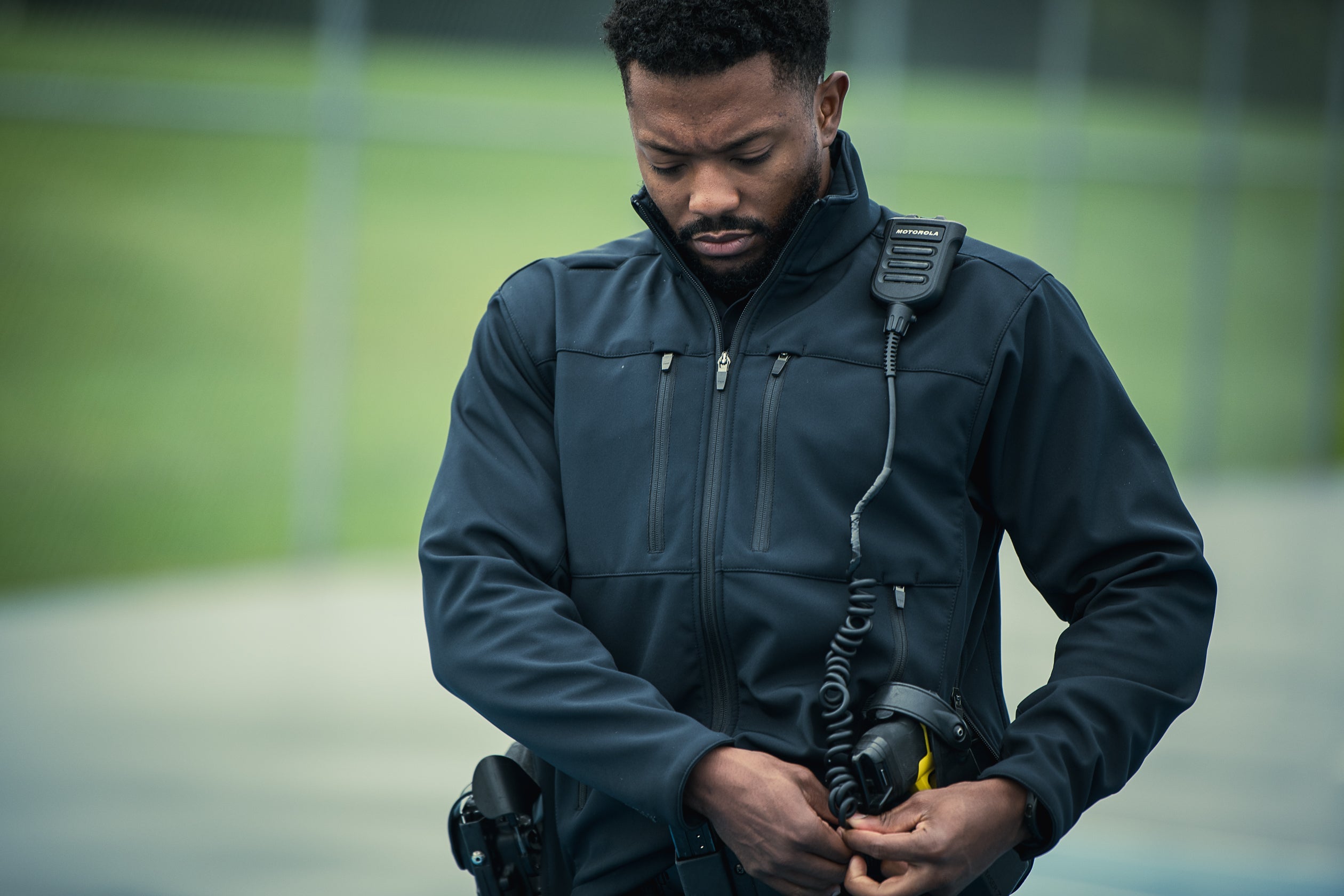 A photograph shows a man in a dark jacket adjusting his Motorola radio outdoors against a blurred green background with hints of a fence and track