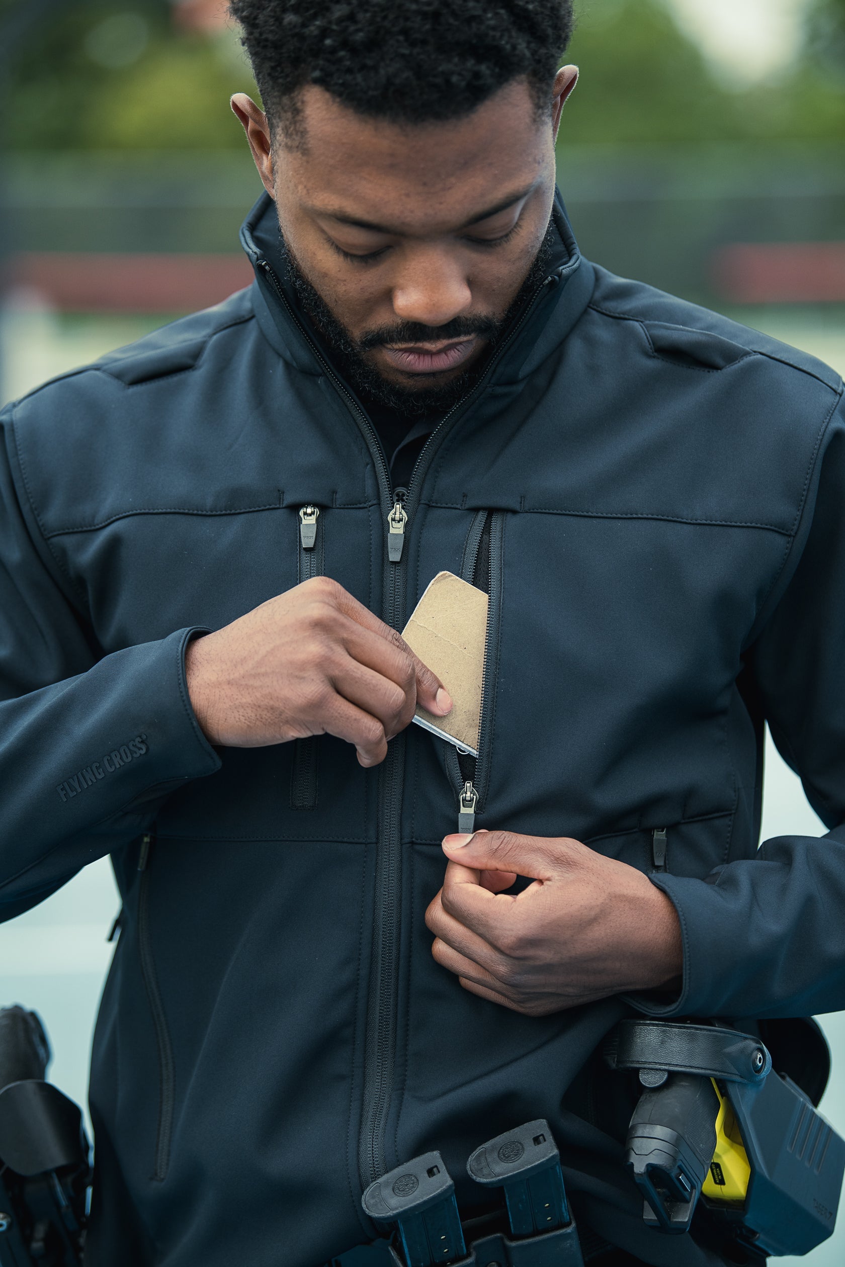 Photography a man wearing a dark Flying Cross softshell jacket with zippered pockets and a tan item inside outdoors near green foliage and a blurred fence showing the jacket's detail and logo