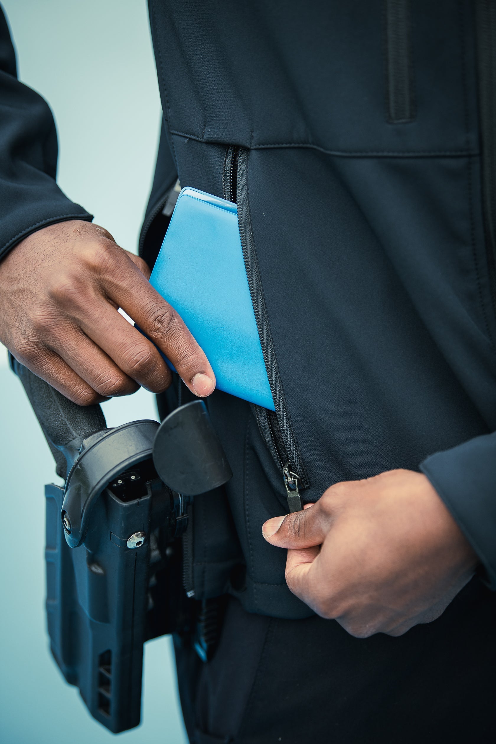 Photograph of a person's hands accessing a bright blue object inside a dark jacket pocket against a light blue background
