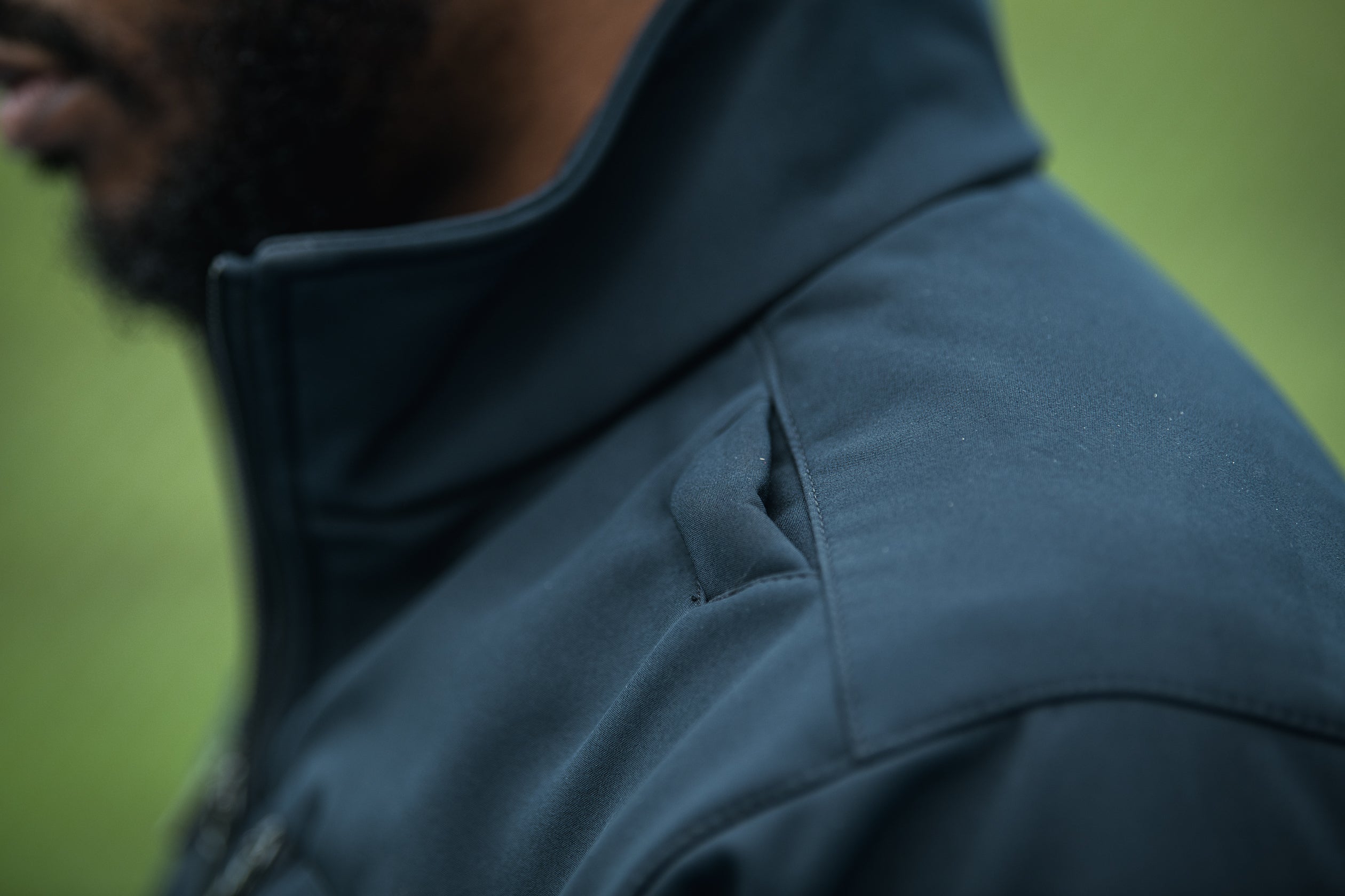 Close-up photograph showing a dark teal softshell jacket with a high collar and subtle shoulder paneling featuring a textured fabric and a zipper detail