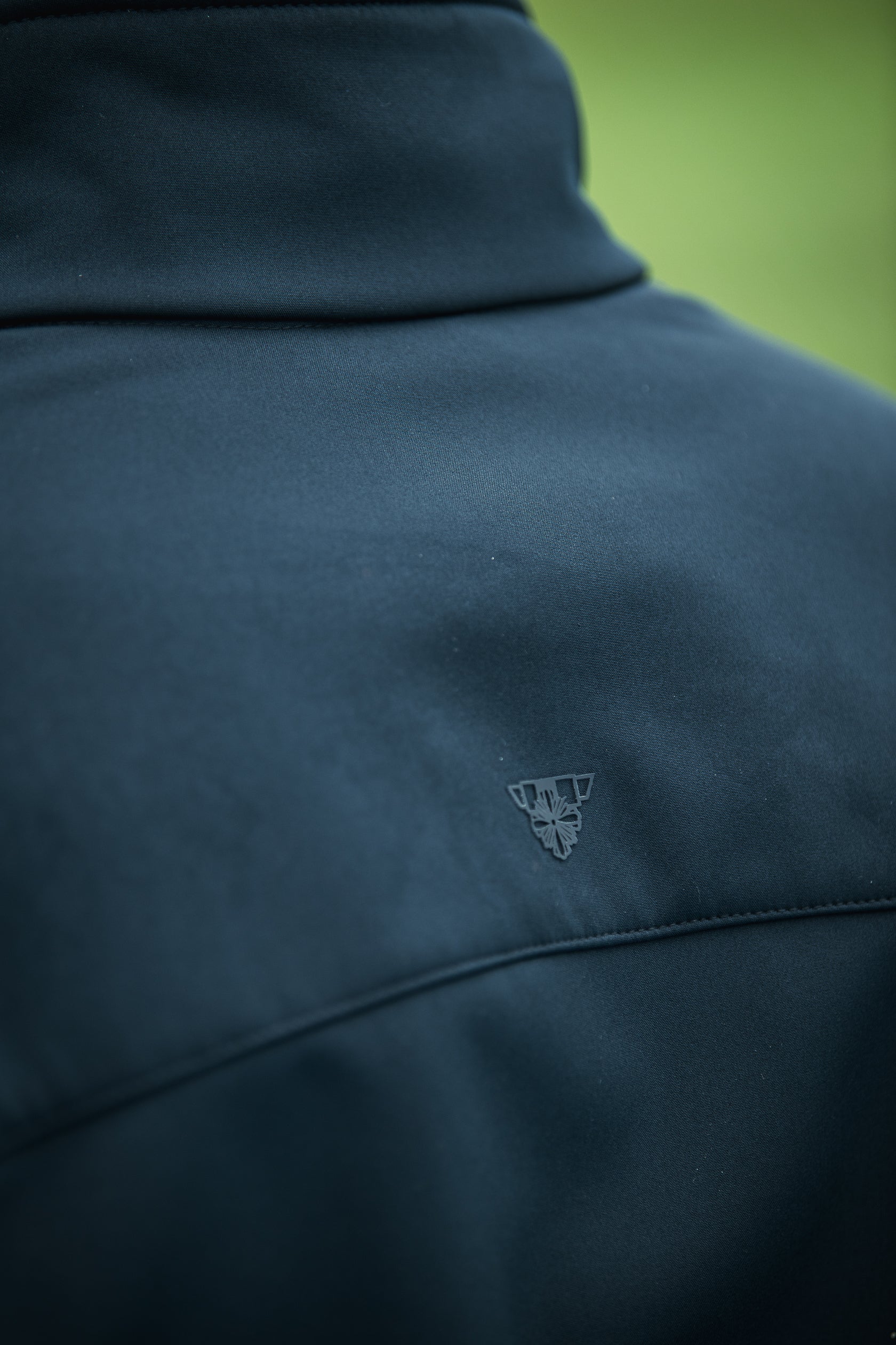 Close-up photograph showing the dark teal textured back of a softshell jacket with a subtle embossed logo featuring a stylized flower within a triangle