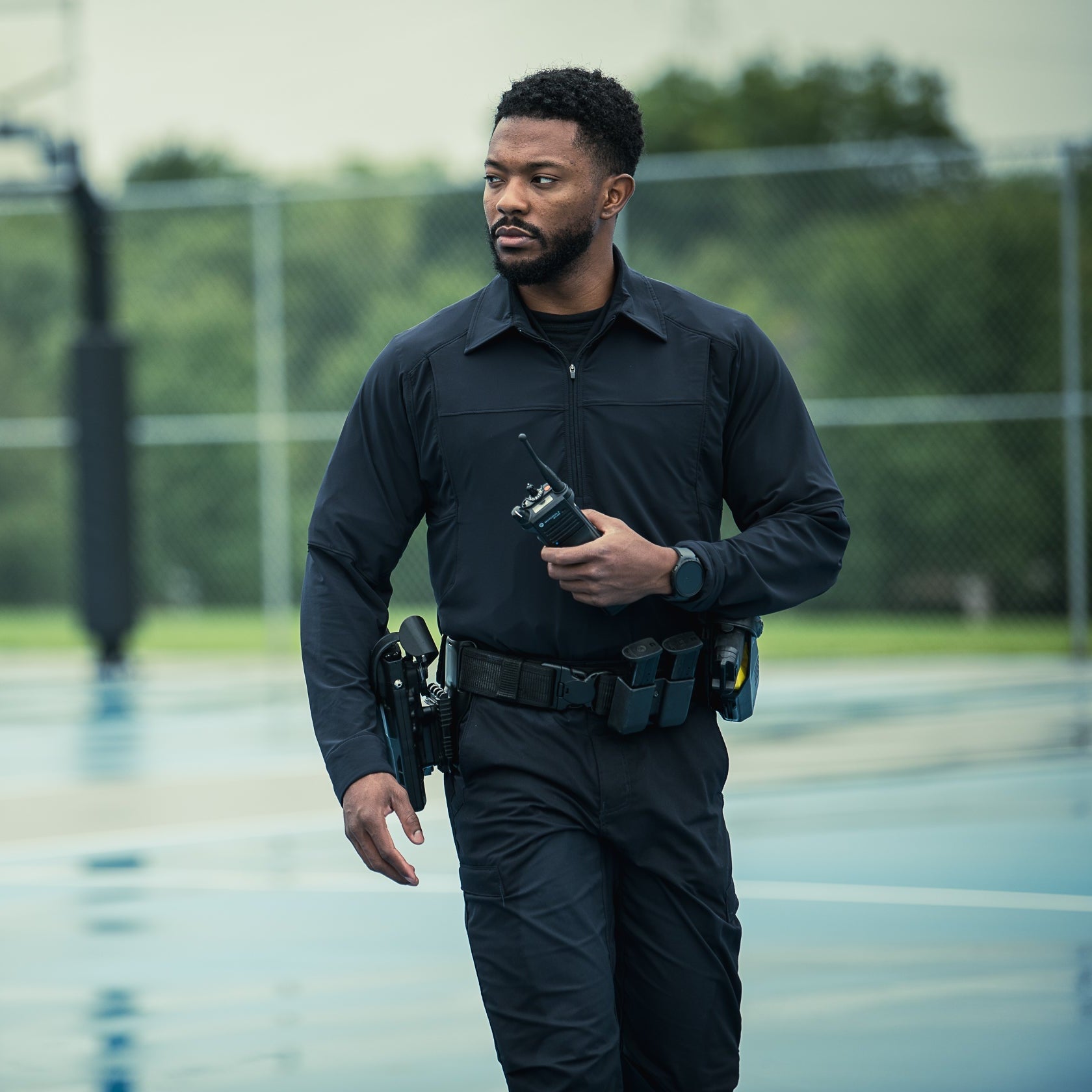 Photograph of a police officer in uniform on a basketball court near a chain-link fence and basketball hoop, holding a radio, with a dark watch and utility belt with multiple pouches.