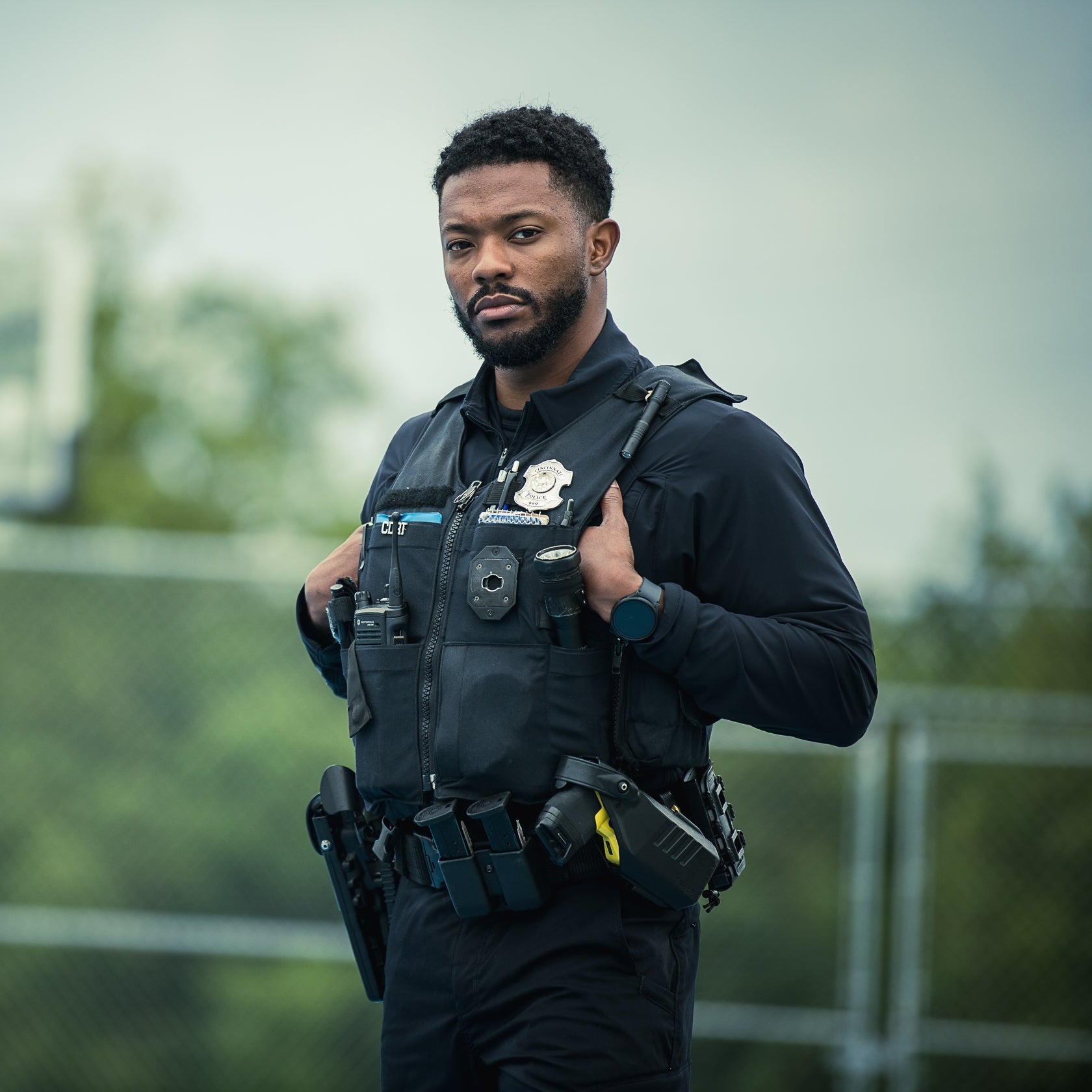 Photograph of a Cincinnati police officer outdoors wearing a black uniform with a body camera, radio, and firearm a serious expression and smartwatch