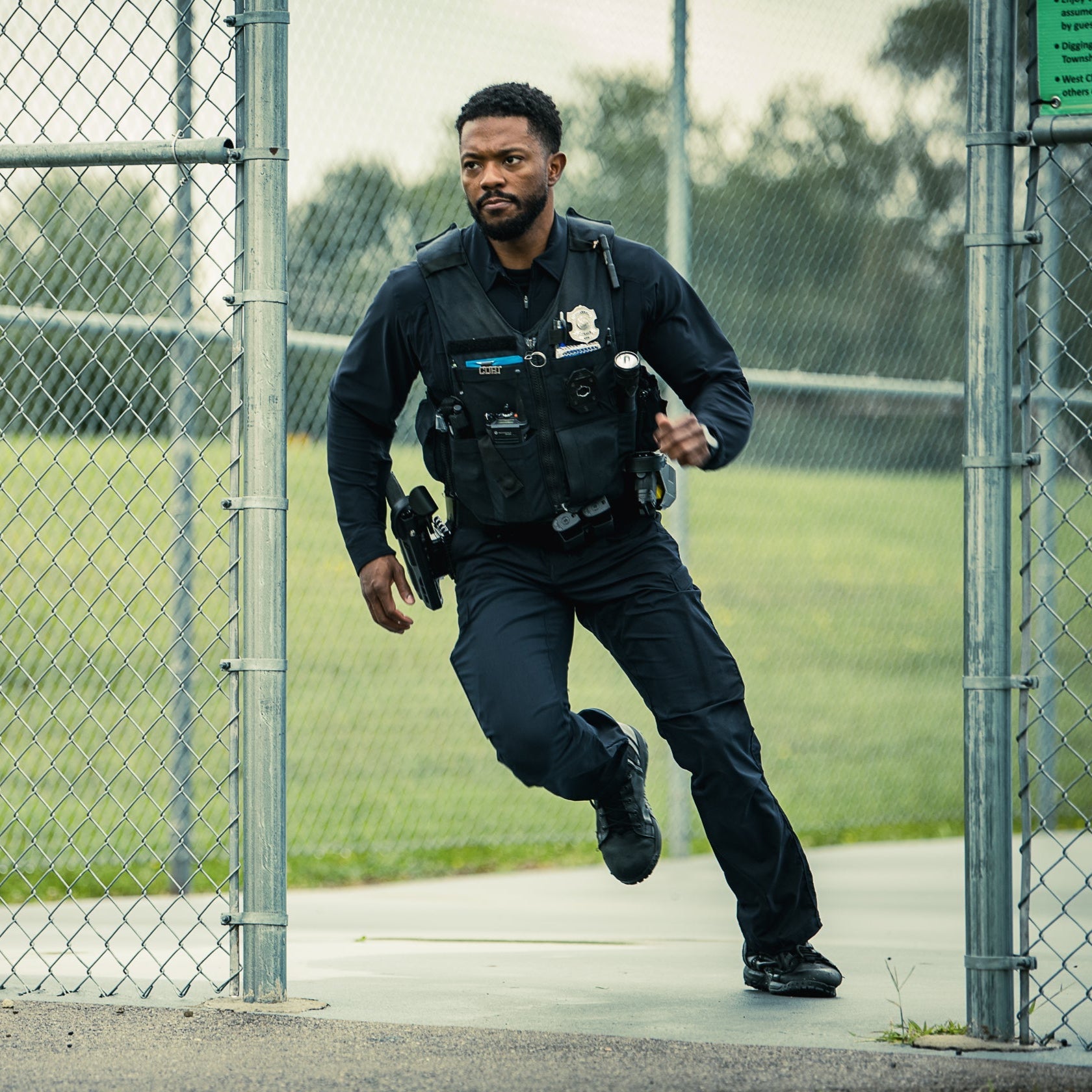Photo of a police officer running through a chain link fence at a park, showcasing his dark uniform, tactical vest with various attachments, and sturdy boots