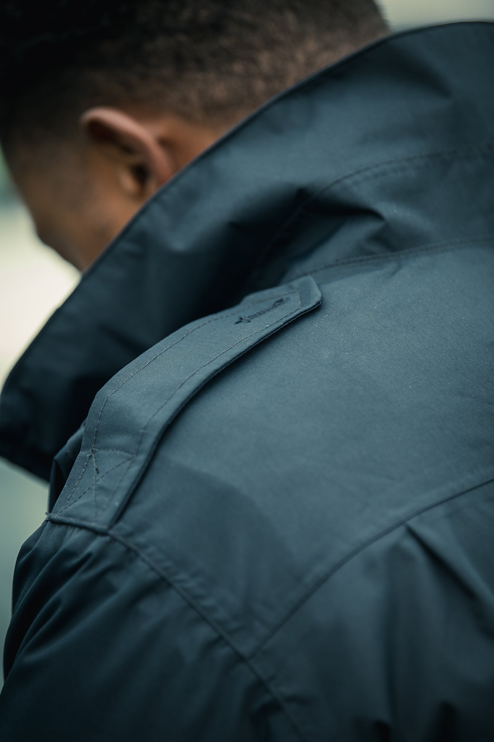 Photographic image of a dark teal jacket showcasing detailed stitching, a noticeable shoulder yoke, and subtle textural variations in the fabric