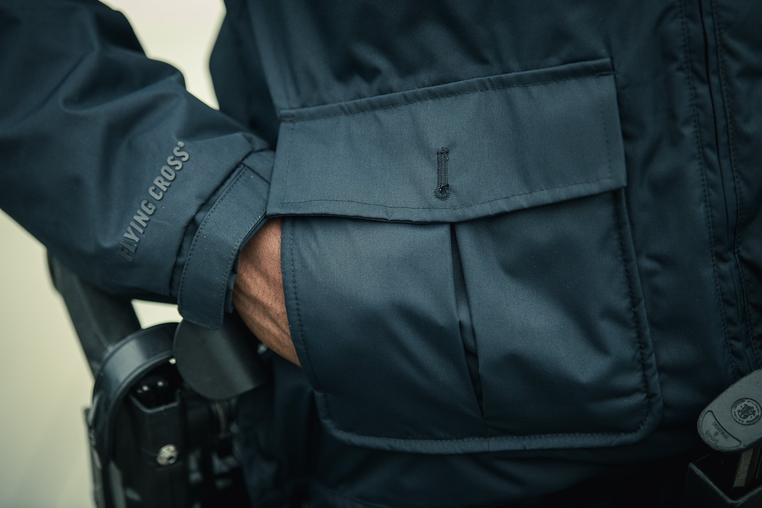 Close-up photograph showing a dark teal Flying Cross jacket with a large front pocket, a dark gray embroidered logo, and subtle stitching details