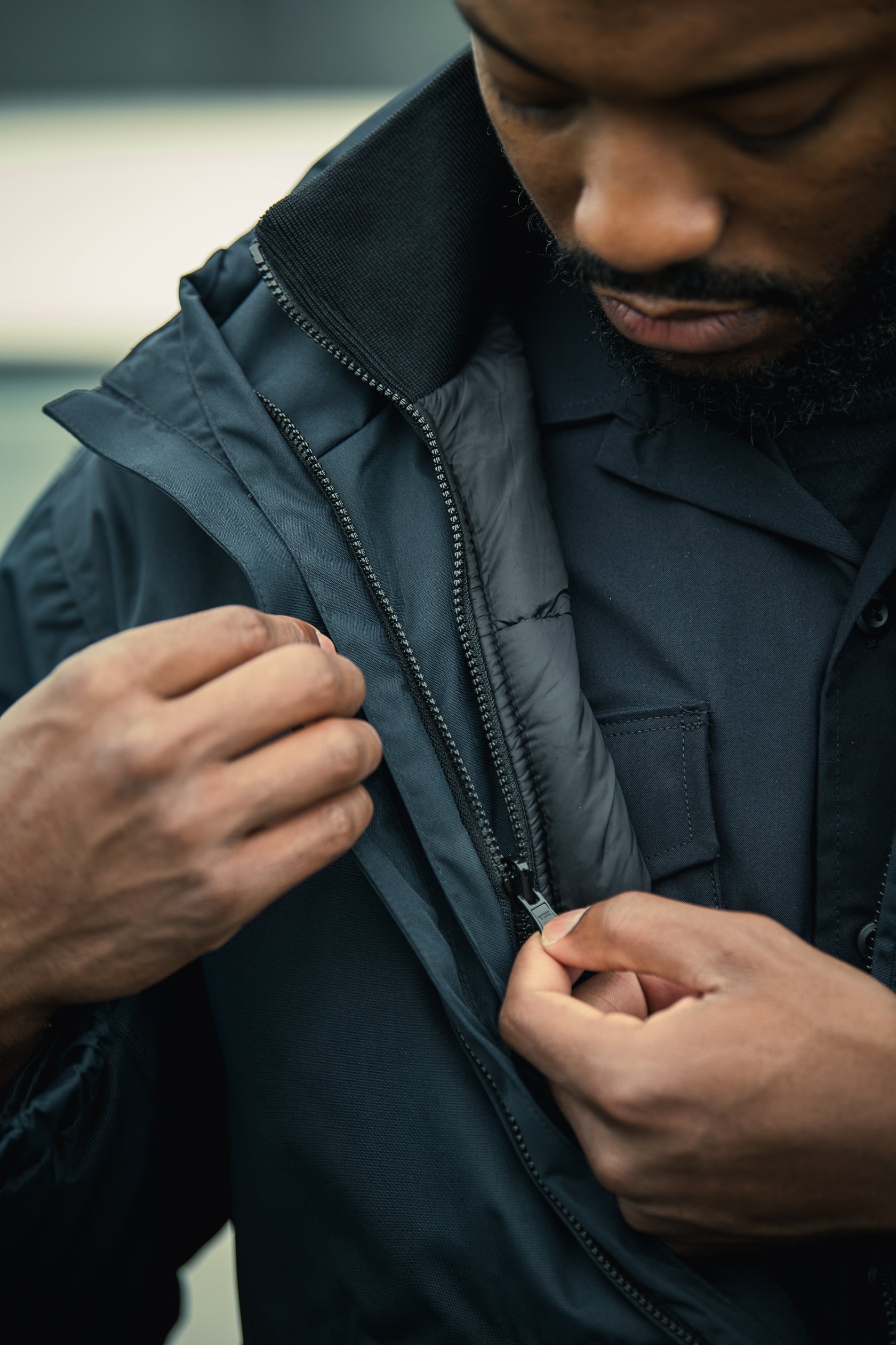 Close-up photograph of a person zipping up a dark teal jacket with a black zipper and ribbed collar showcasing the textured fabric and subtle sheen of the garment.