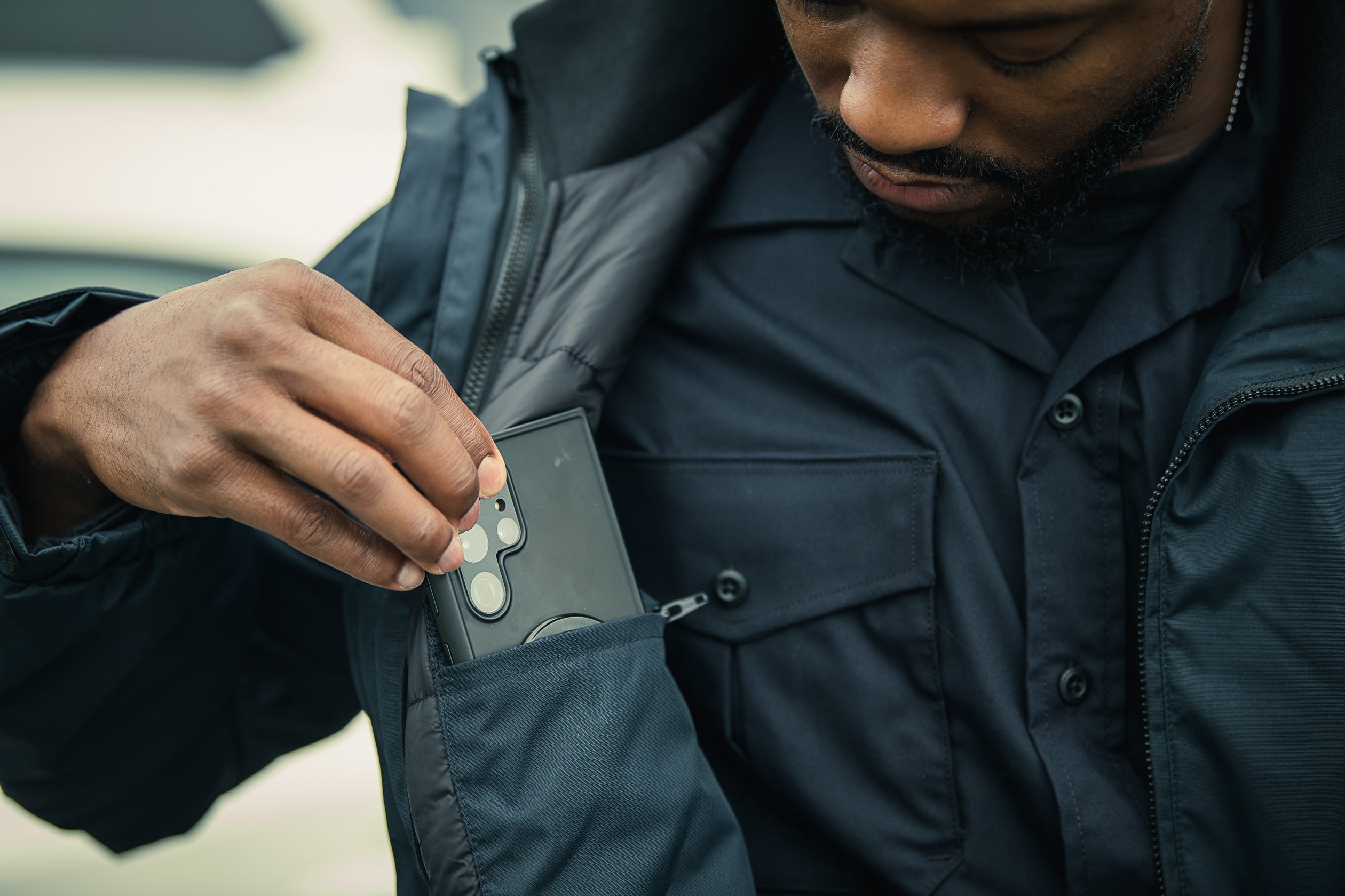 Close-up photography of a person placing a black smartphone with a distinctive multi-lens camera system into a dark jacket pocket, showcasing the device's sleek design and textured fabric of the clothing