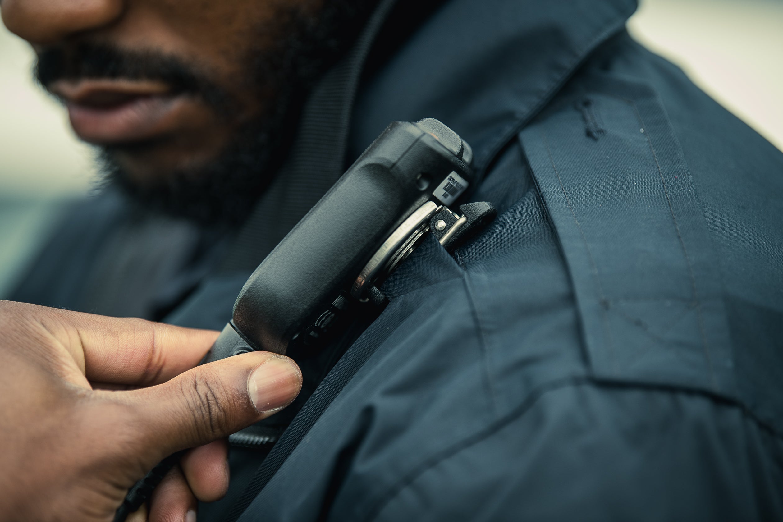 A photograph shows a close-up of a person's hand adjusting a black radio on their dark blue uniform showcasing a silver clasp and a barcode detail the image lacks discernible landmarks or colorful elements