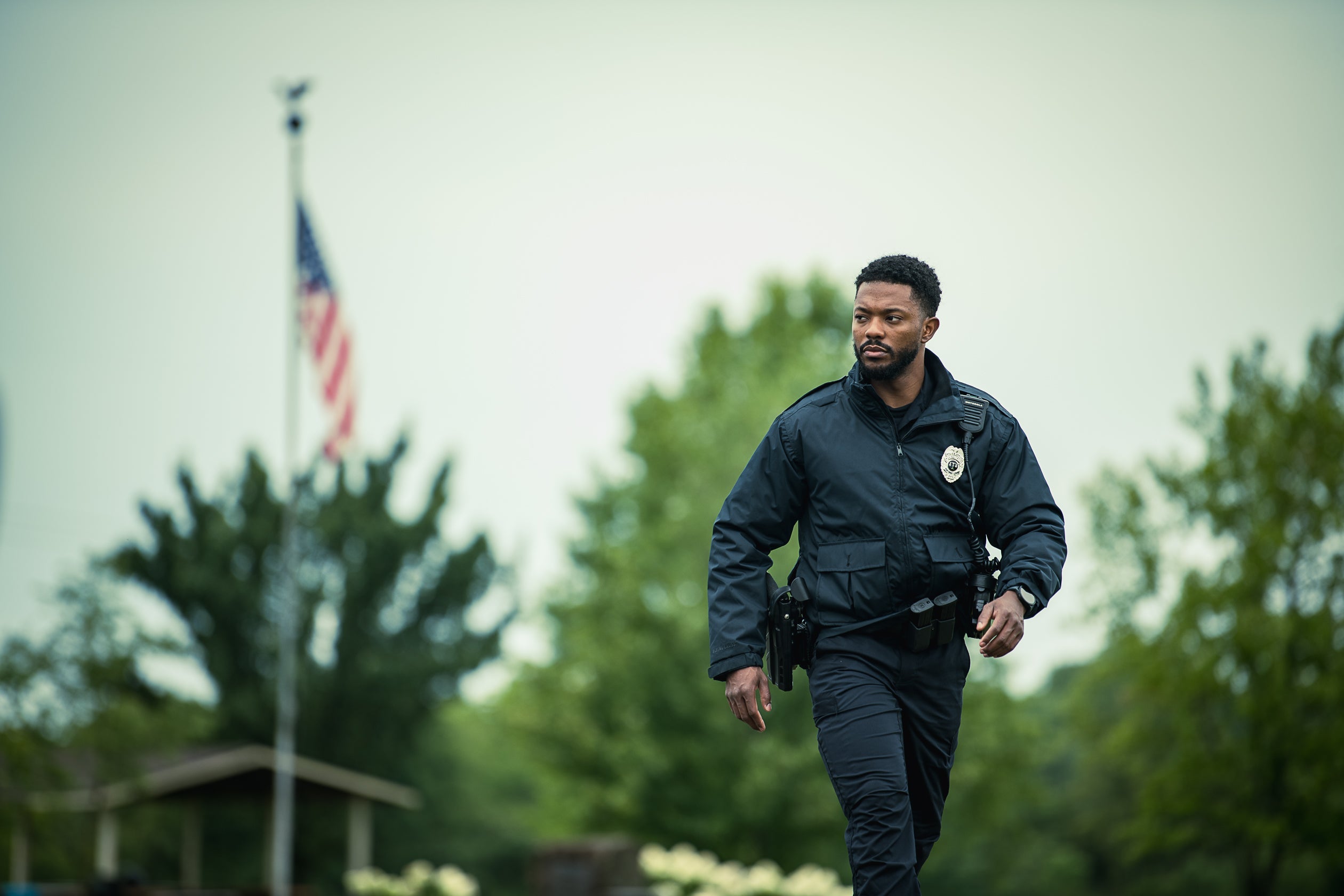 Photograph of a police officer in uniform walking outdoors near an American flag a park pavilion and lush green trees his dark jacket and badge are visible along with a smartwatch