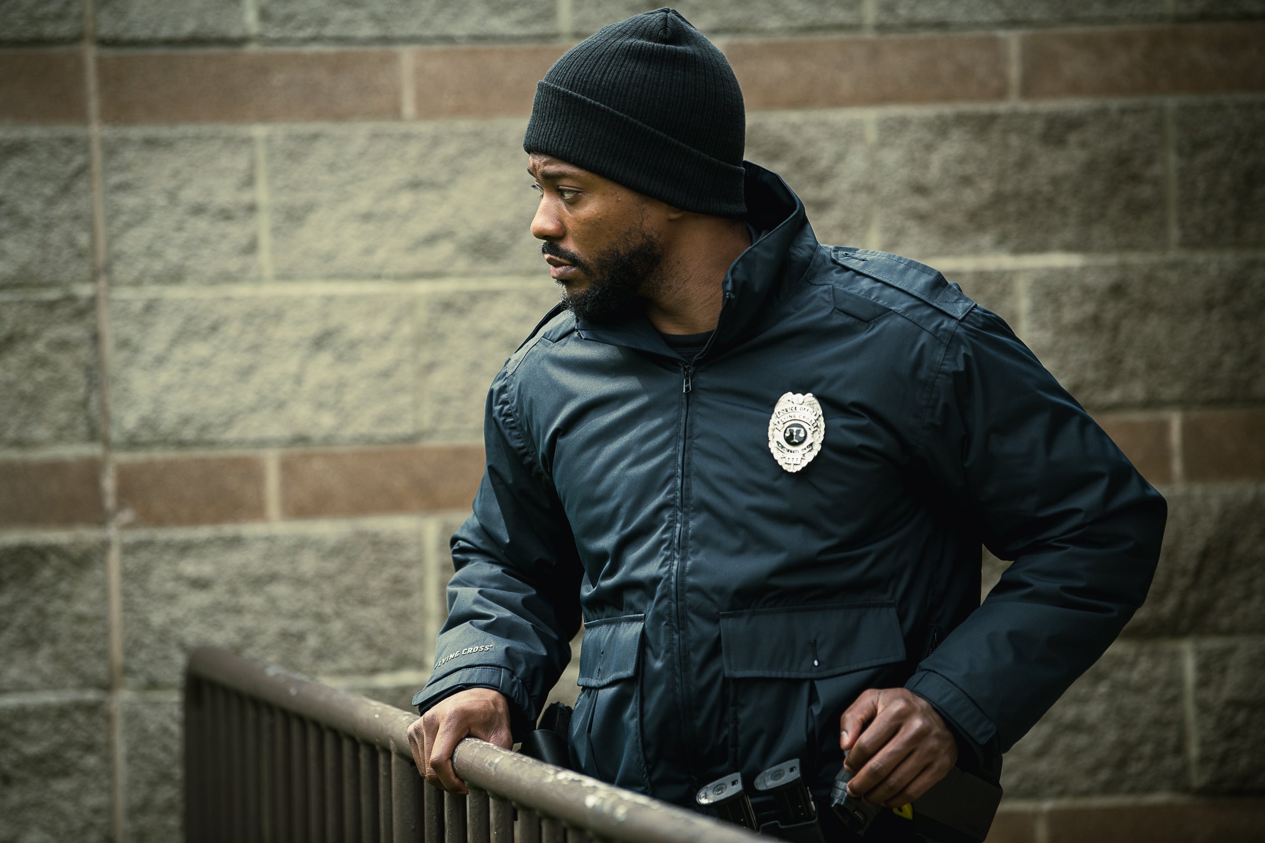 Photograph of a Black police officer in a dark jacket and black beanie standing by a grey brick wall with brown accents and a dark metal railing holding a radio