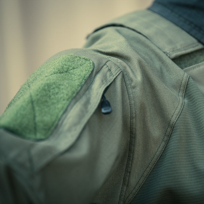 Close-up photograph of a military-style olive green jacket showing a Velcro patch, zippered pocket, and textured fabric detail