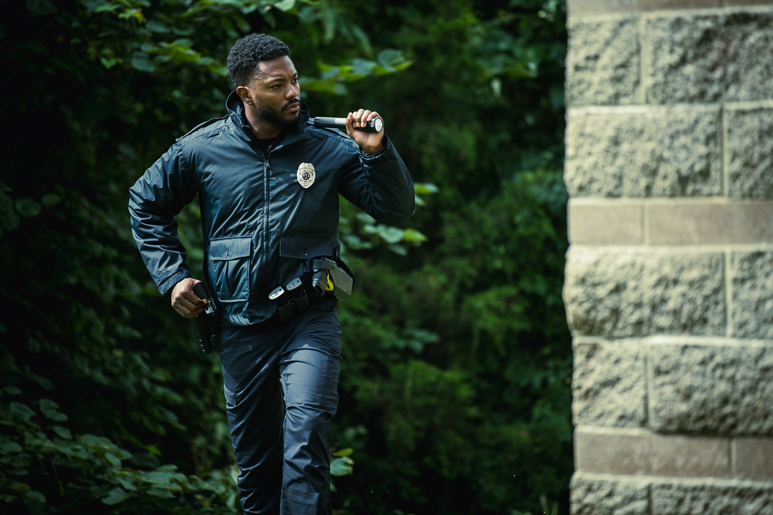 Photograph of a police officer in dark uniform, carrying a flashlight and firearm, running through a lush green park near a textured stone wall.