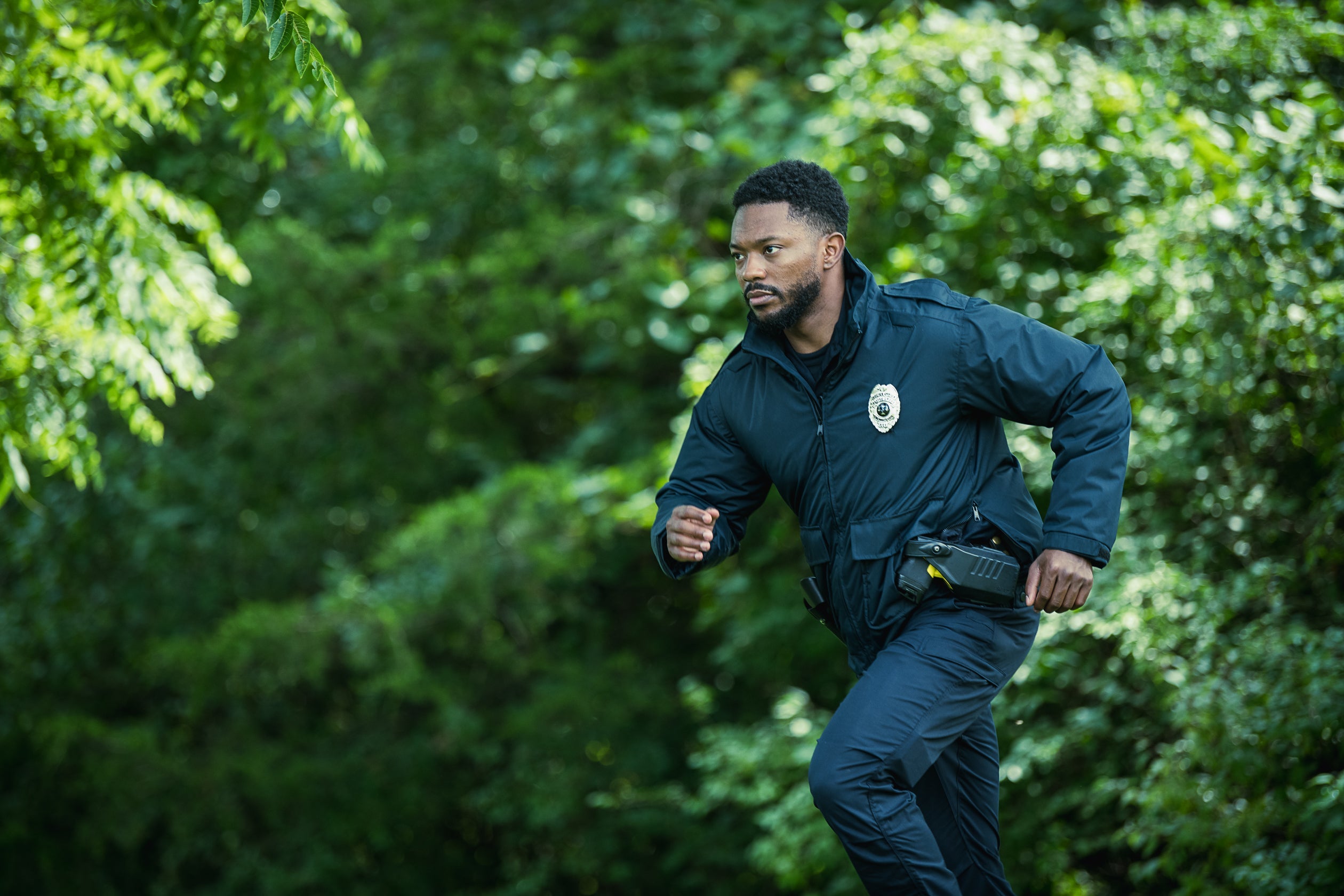 Photograph of a police officer running through a lush green forest, his dark blue uniform, shiny badge, and a partially visible yellow detail on his holster stand out against the vibrant foliage