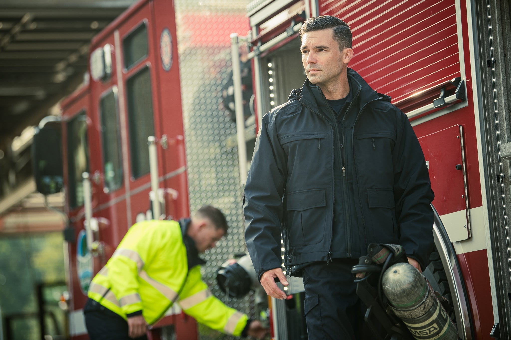 Photograph firefighter in dark jacket holding oxygen tank near red firetruck with visible emblem and a bright lime green jacket in background