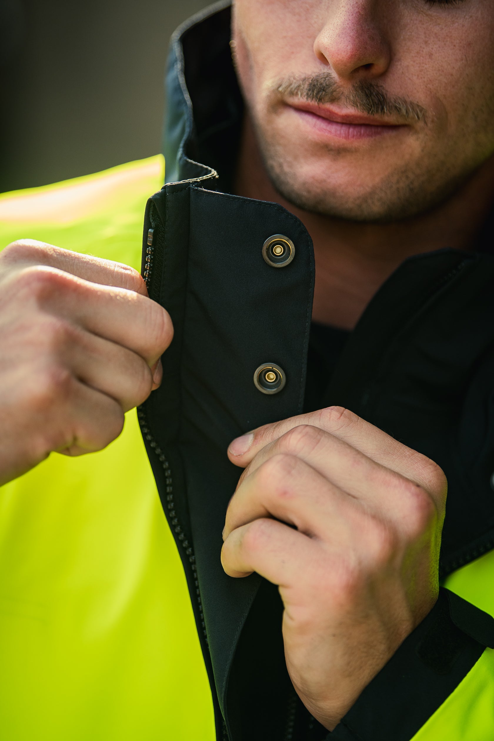 Close-up photograph showing a person fastening a black jacket with brass buttons over a bright yellow shirt