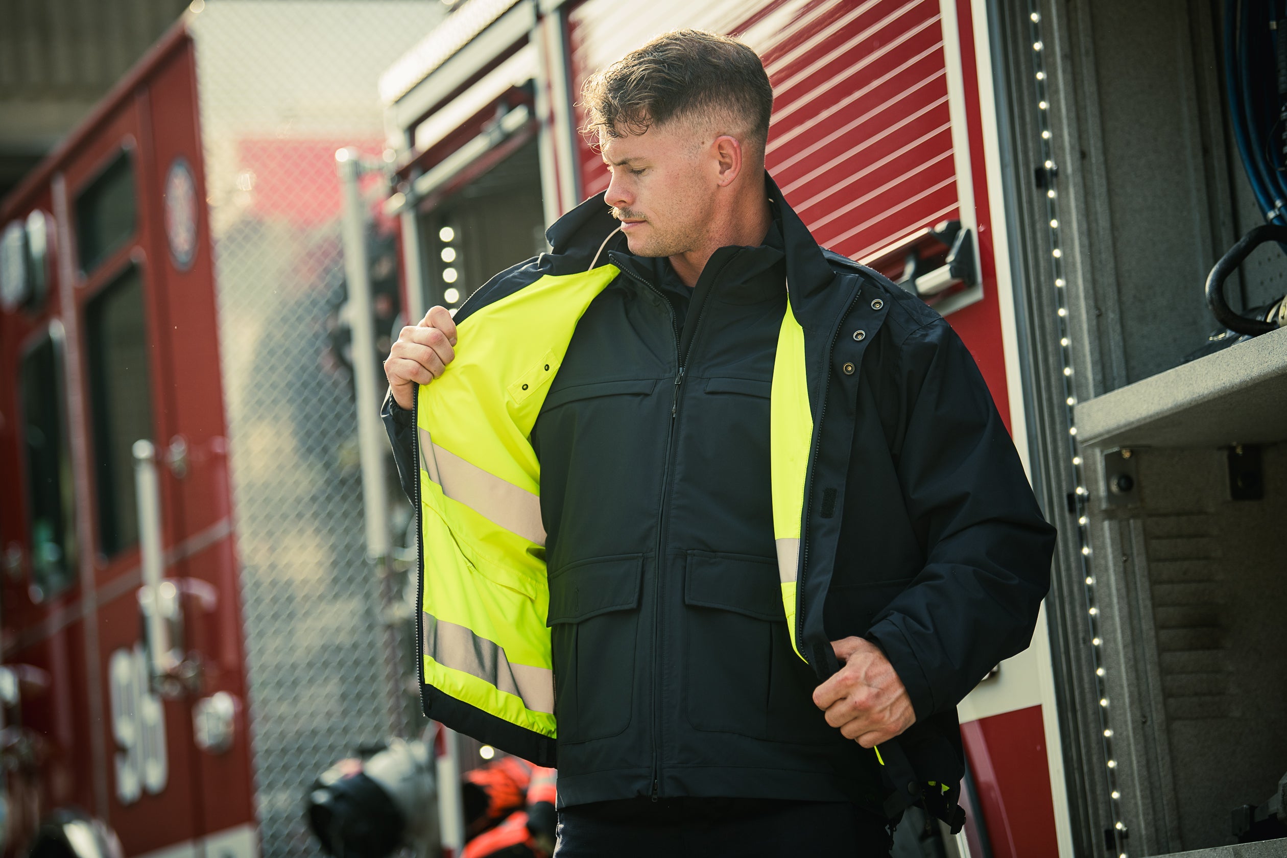 Photograph showing a man in a black jacket with a bright yellow interior and reflective strips standing in front of a red firetruck with visible number and LED lights