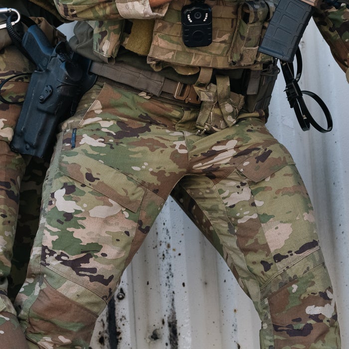 Photograph of camouflage military pants against a weathered metal wall showing a handgun holster, body camera, and multiple cargo pockets with varied shades of green and brown