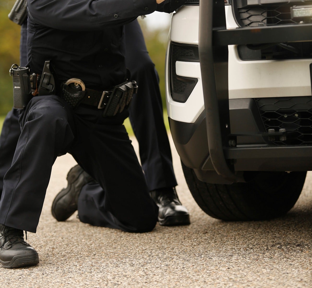 Photograph of two police officers kneeling near a white police vehicle with a black bull bar, showcasing their tactical gear including handcuffs and radios, on a paved road.