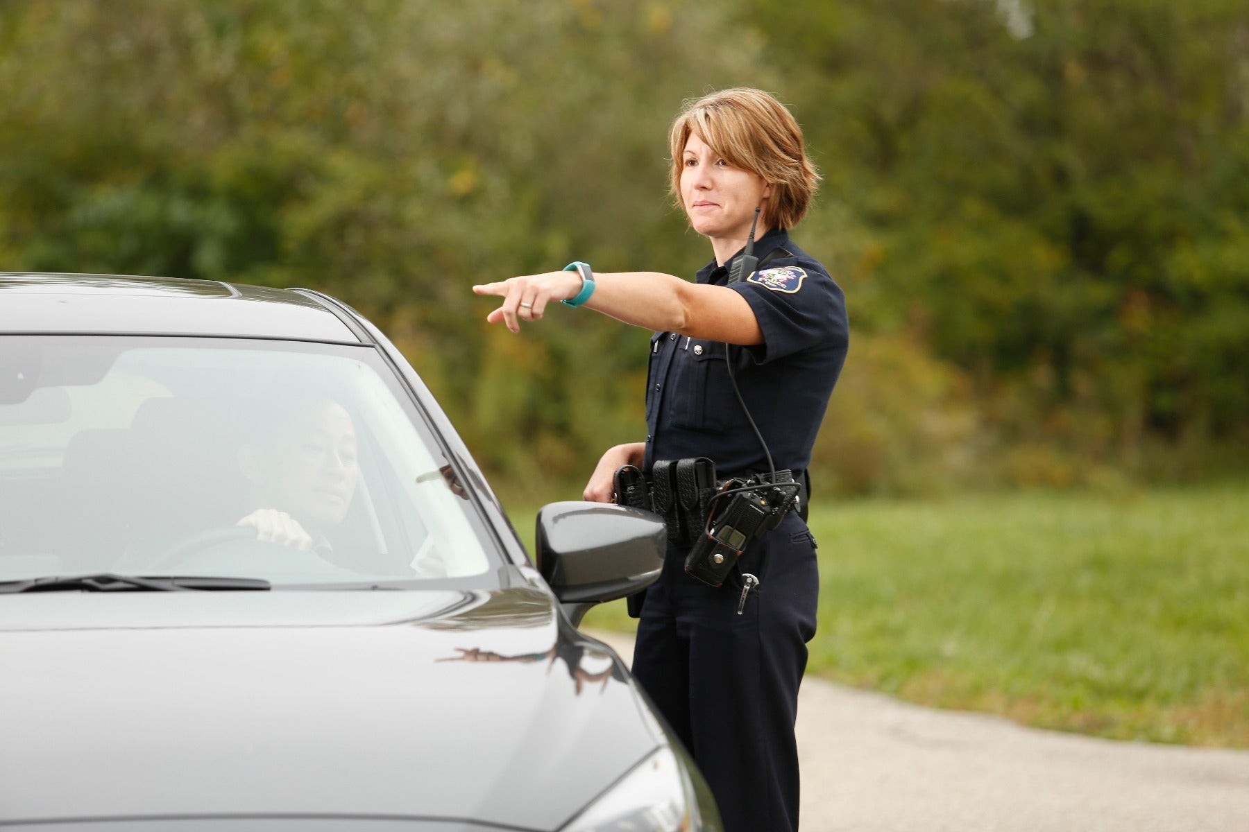 Photograph A female police officer directs a driver during a roadside stop in a verdant setting with a teal fitness tracker, black police radio, and a dark-colored vehicle